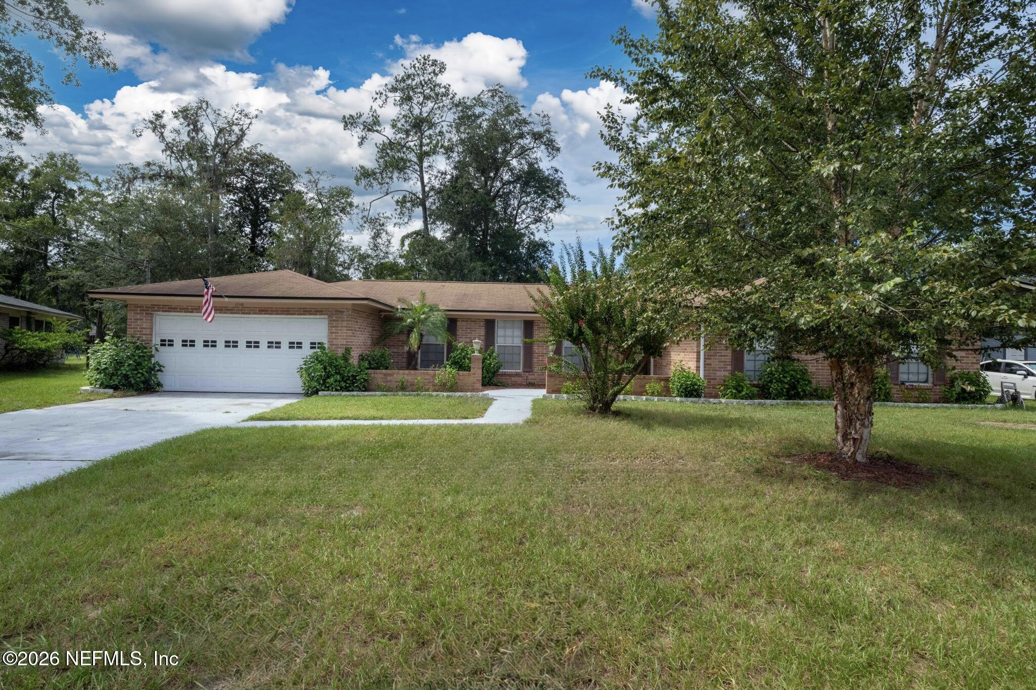 Single-story brick house with a white double garage, broad lawn, mature trees, and an American flag.