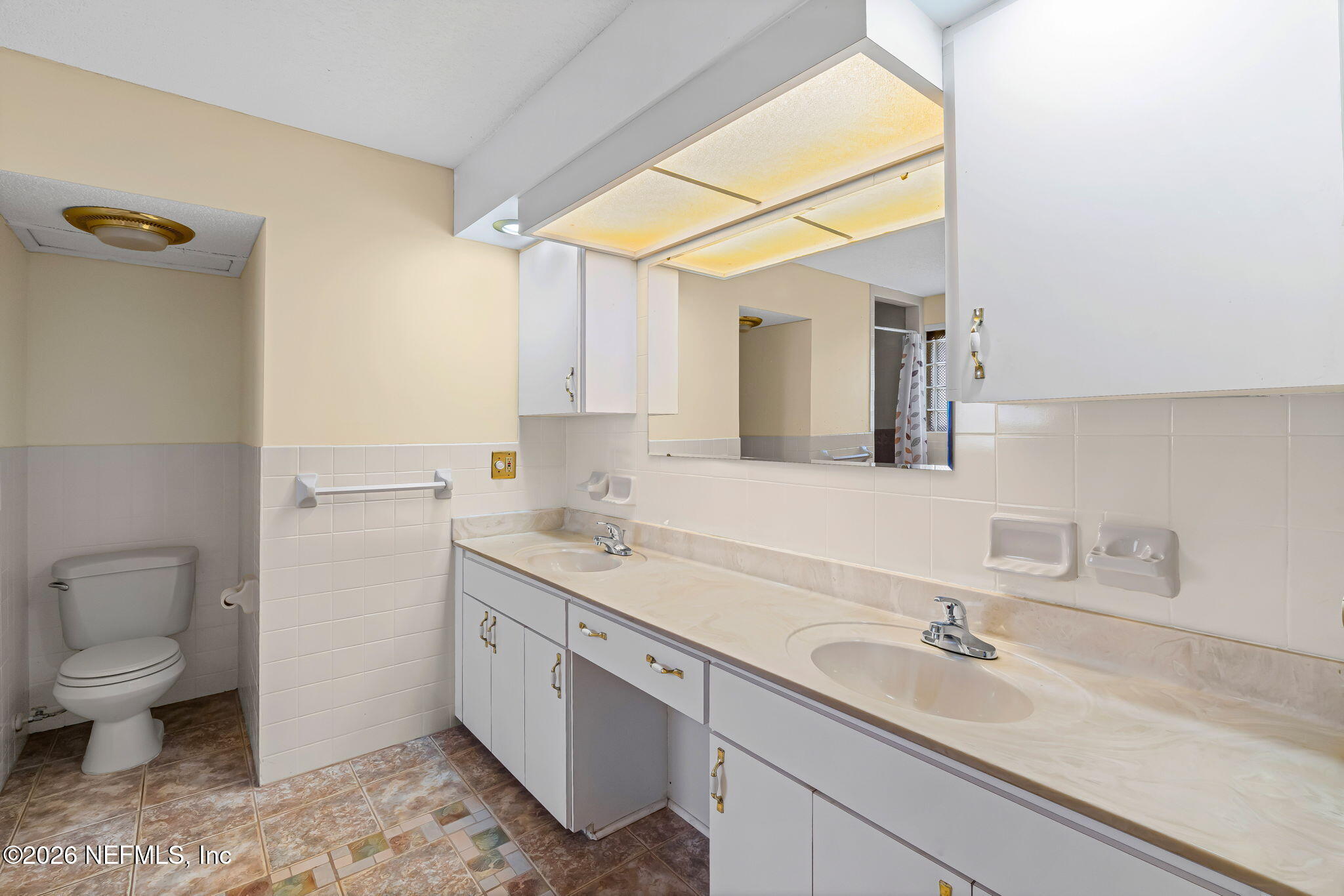 Bathroom with a long double-sink vanity, large mirror, tiled walls, and a toilet alcove.