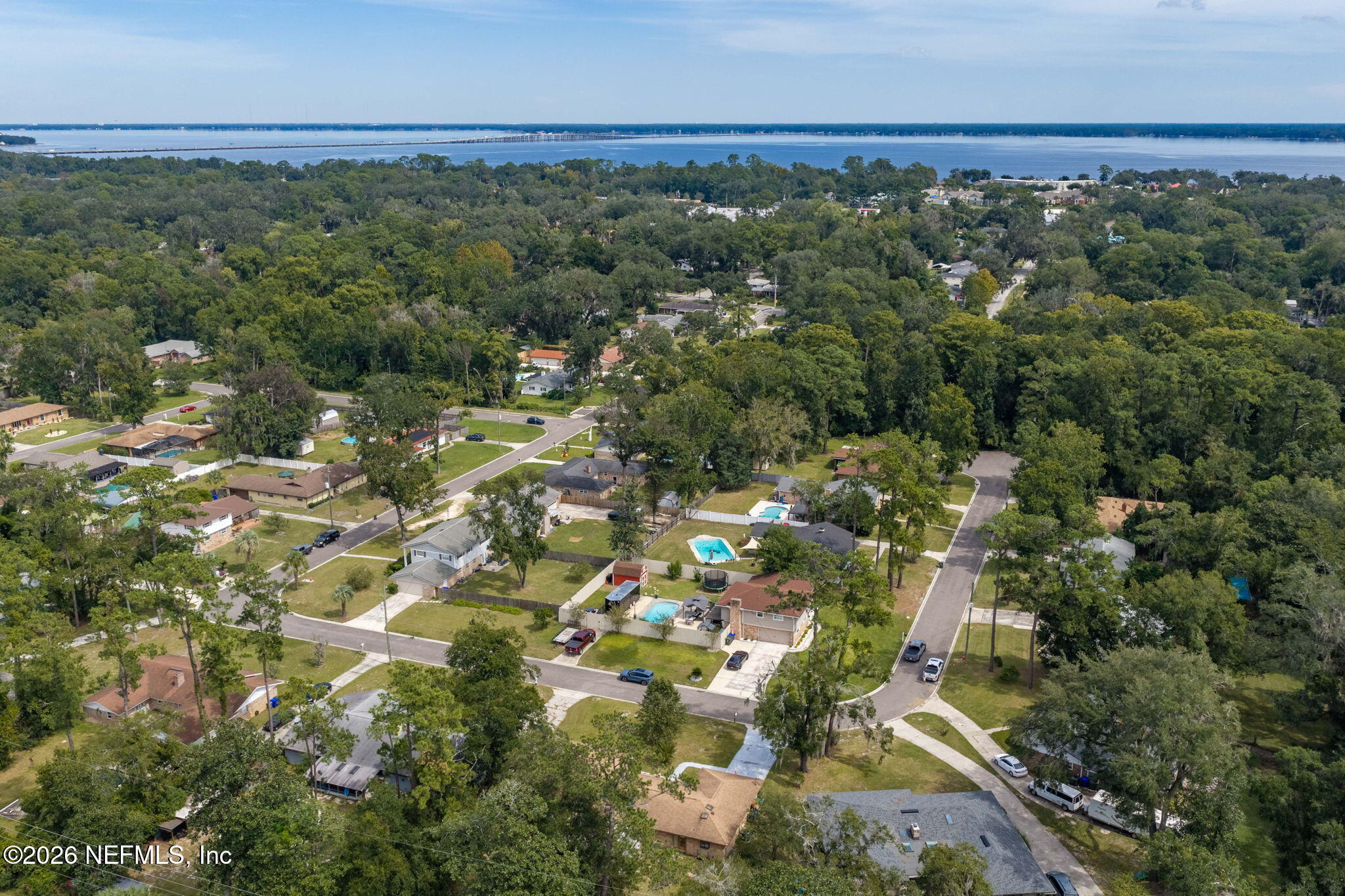 Aerial view of a leafy neighborhood with backyard pools and distant water and bridge.