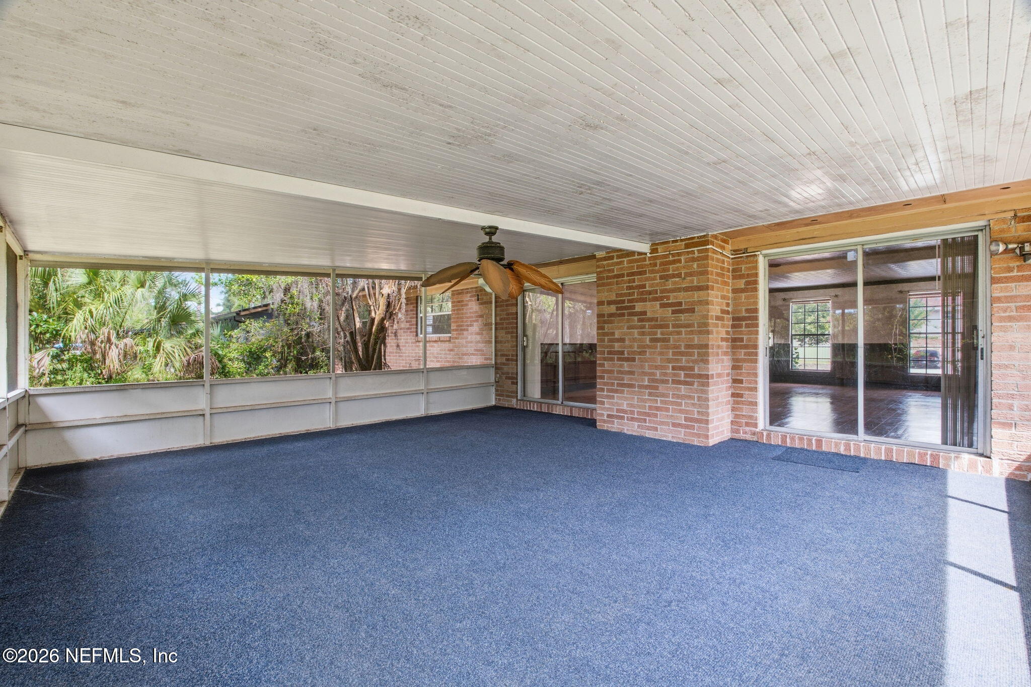 Screened-in porch with blue carpet, a ceiling fan, brick walls, and views of leafy greenery.