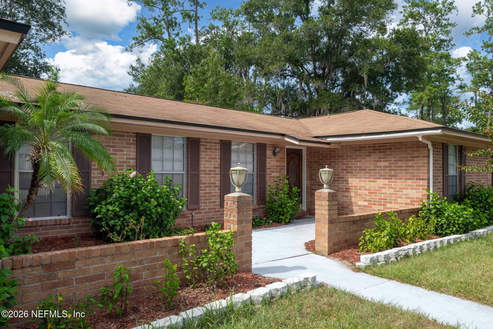 Single-story brick home with dark shutters, landscaped front beds, and a walkway framed by lantern-topped pillars.