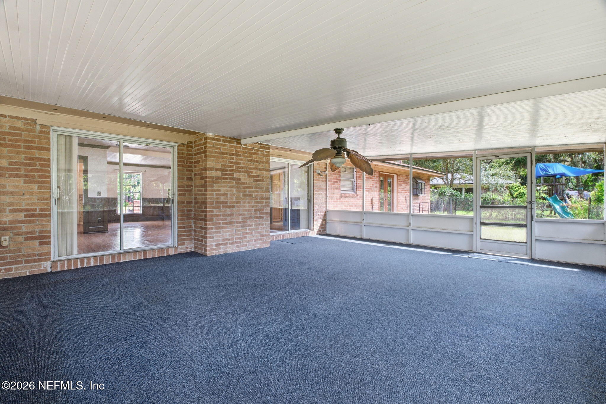Enclosed porch with blue carpet, brick walls, a ceiling fan, and windows overlooking the backyard.