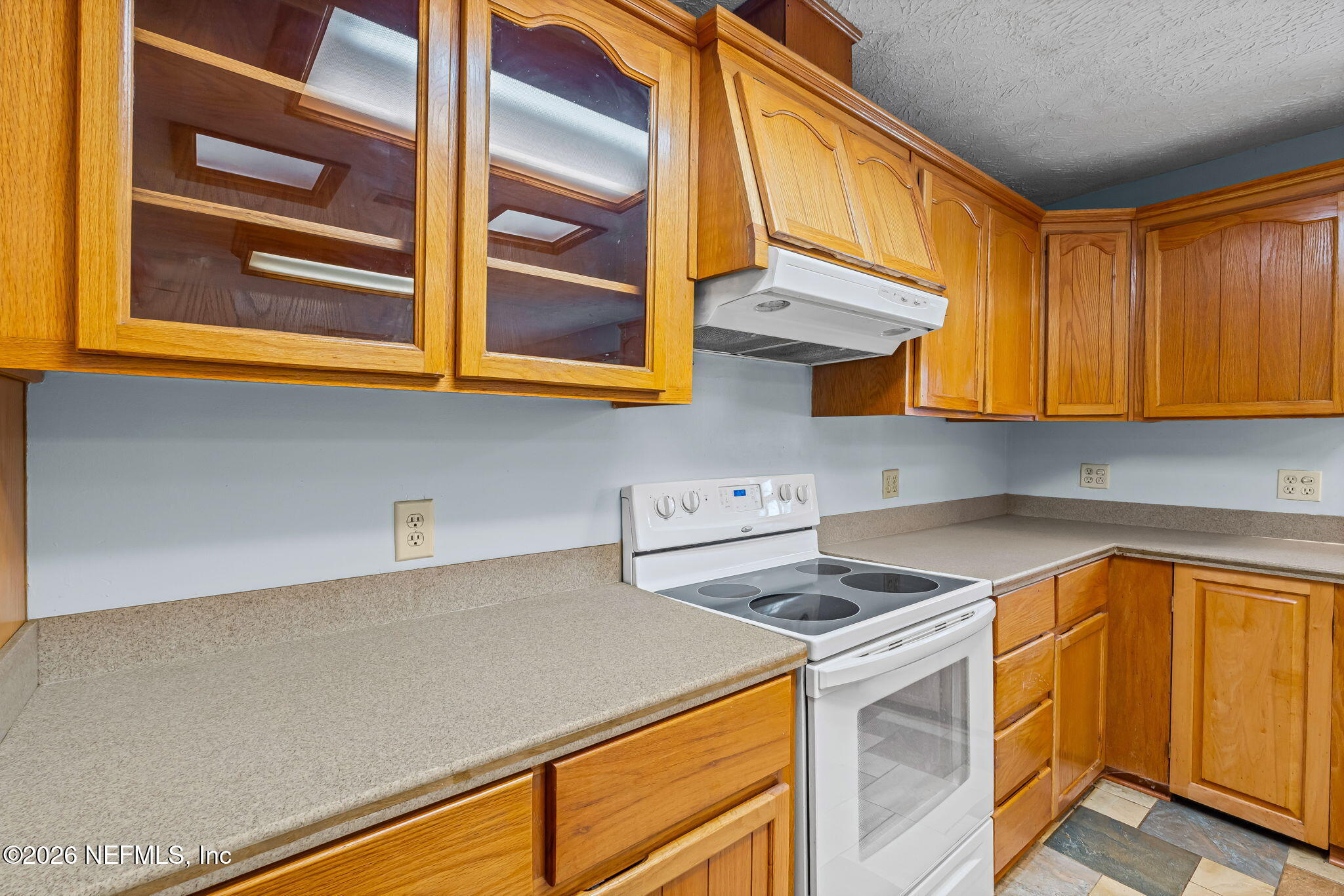 Kitchen with honey-colored wood cabinets, glass-front uppers, beige counters, and a white electric range.