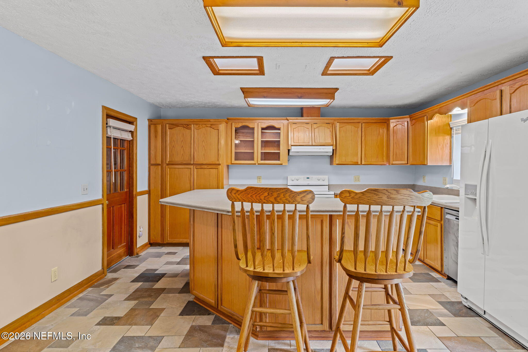 Kitchen with honey-toned wood cabinetry, a central island, two wooden stools, and multicolored tile flooring.