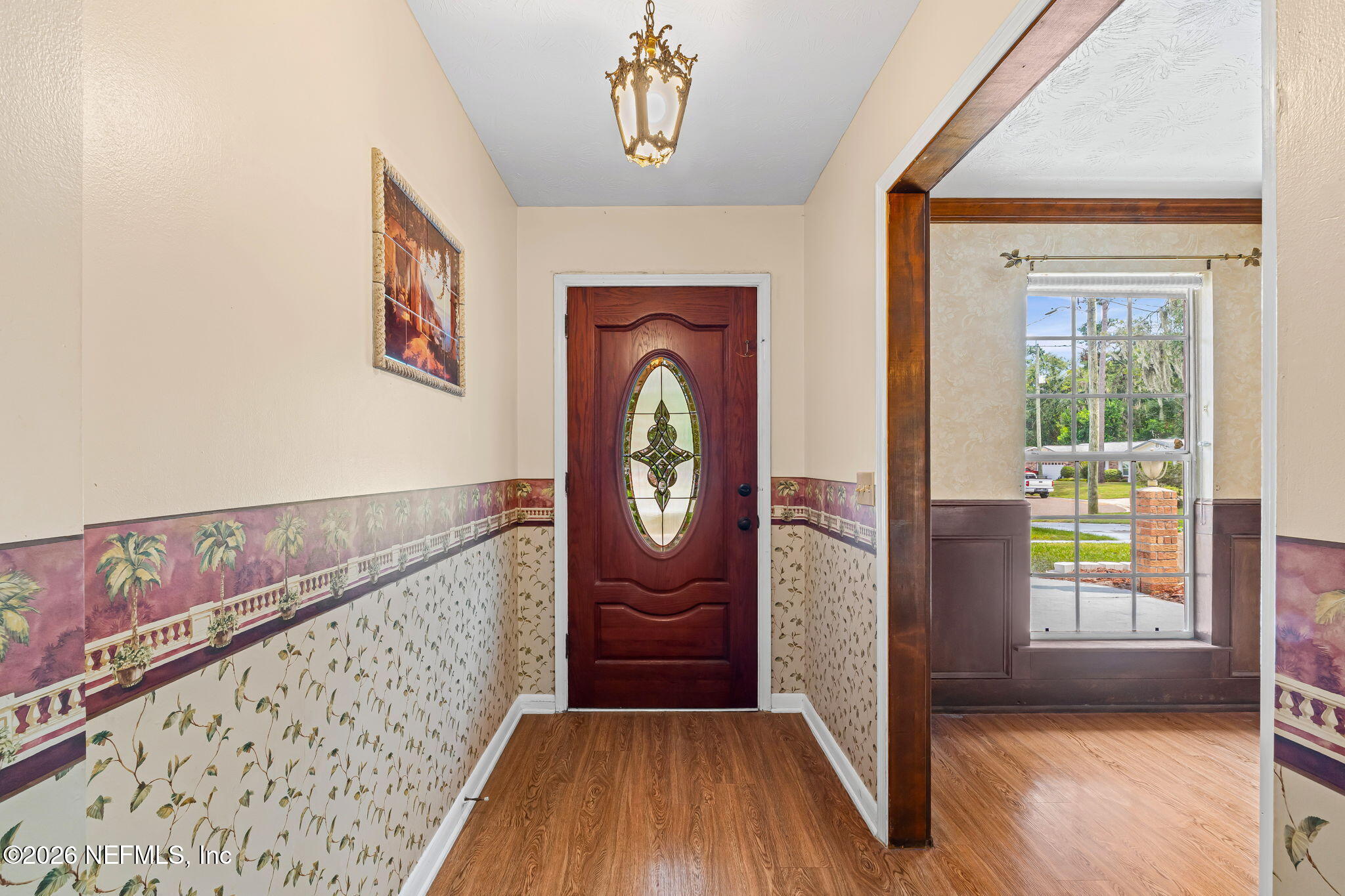 Entry foyer with a stained-glass front door, patterned wallpaper, wood flooring, and a wide opening to a windowed room.