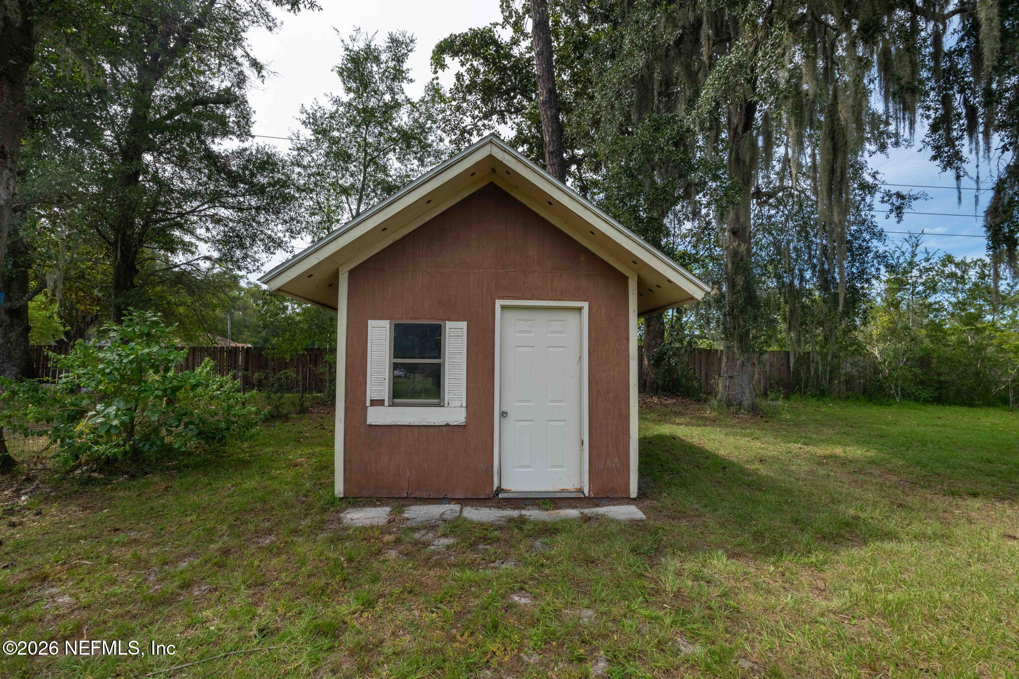 Small brown outbuilding with white trim, a white door, and a shuttered window in a tree-lined yard.