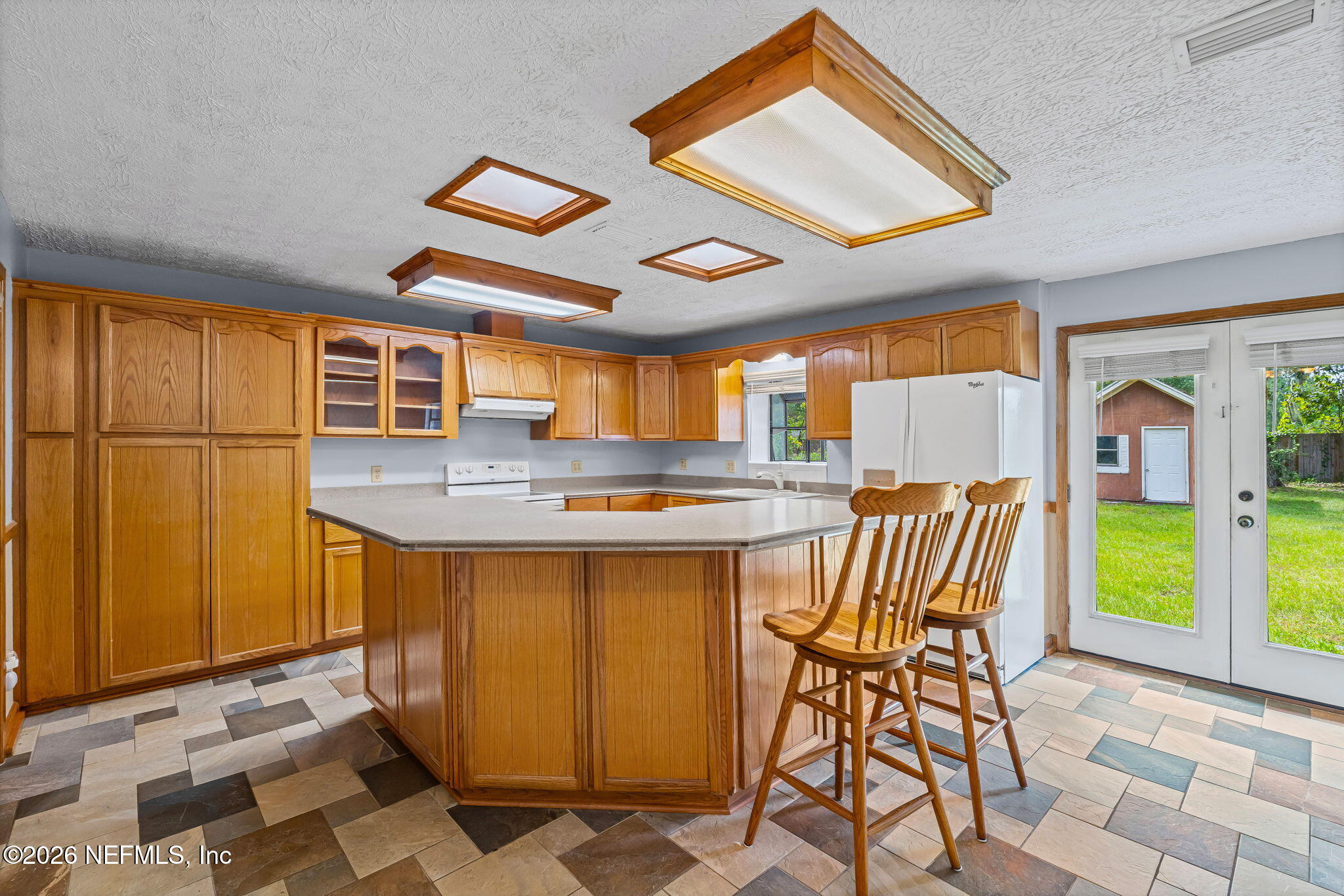 Kitchen with honey-toned wood cabinetry, a large peninsula, two bar stools, and glass doors to the backyard.