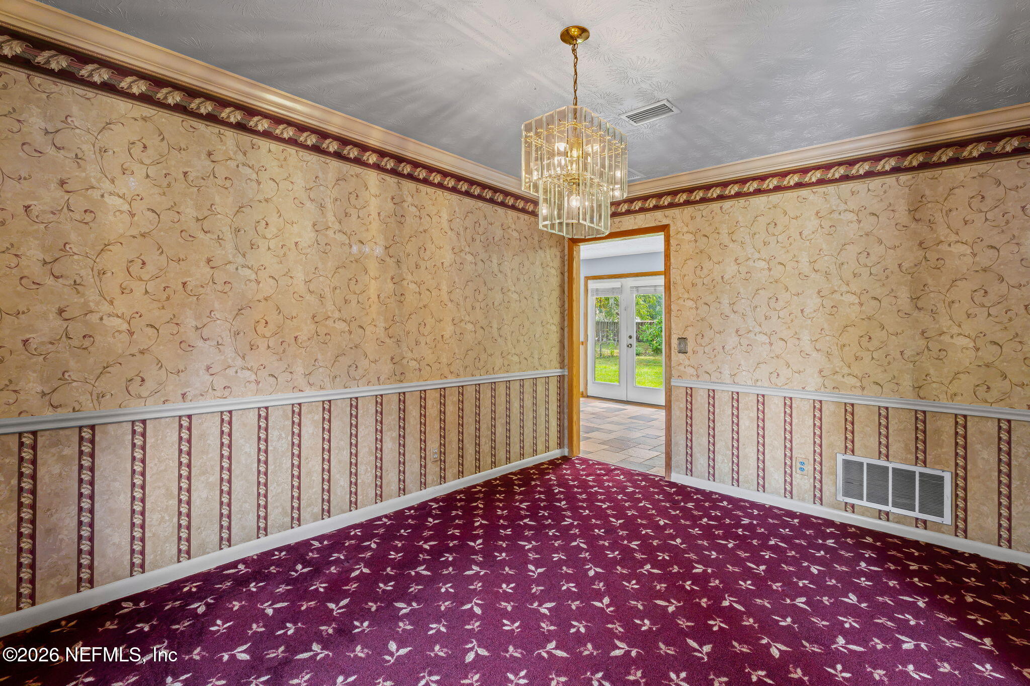 Empty dining room with burgundy floral carpet, ornate wallpaper, and a crystal chandelier.