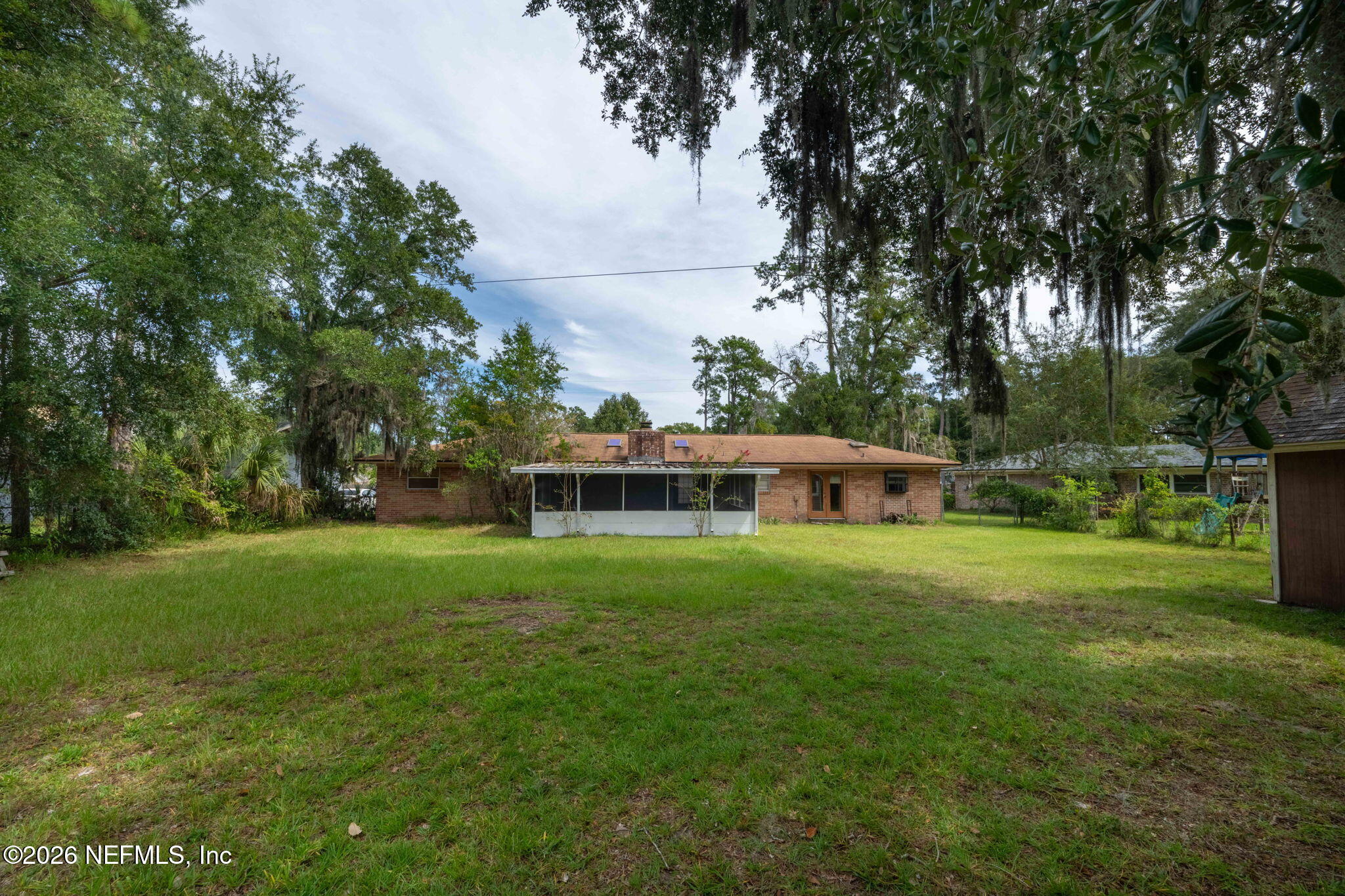Rear of a brick house with a screened porch, spacious grassy yard, and mature trees.
