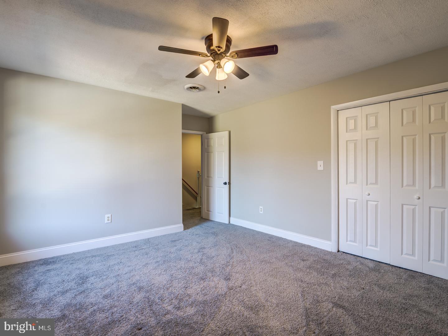 Empty carpeted bedroom with a ceiling fan, white closet doors, and an open doorway.