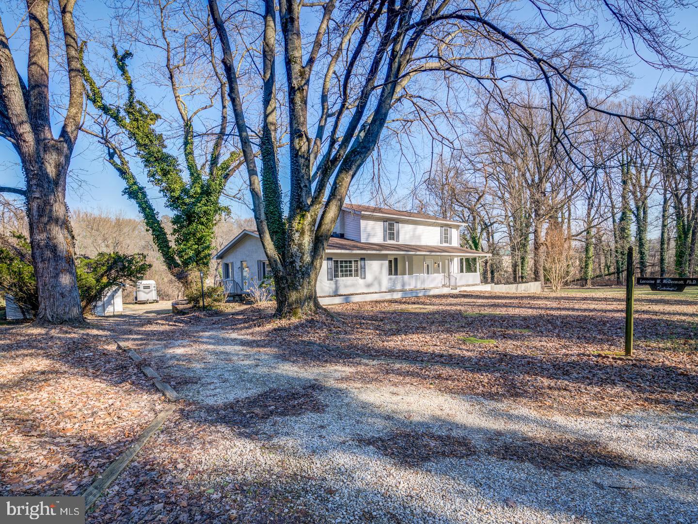 White two-story house with a covered porch, framed by mature leafless trees and a gravel driveway.