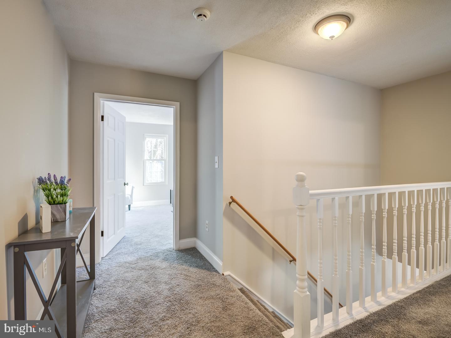 Carpeted upstairs landing with a white stair railing and open doorway to a bright room.
