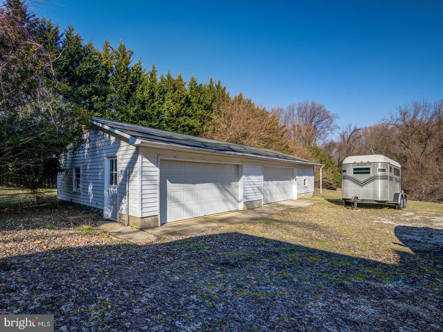 Detached white two-door garage beside a gravel driveway, with a silver horse trailer parked nearby.