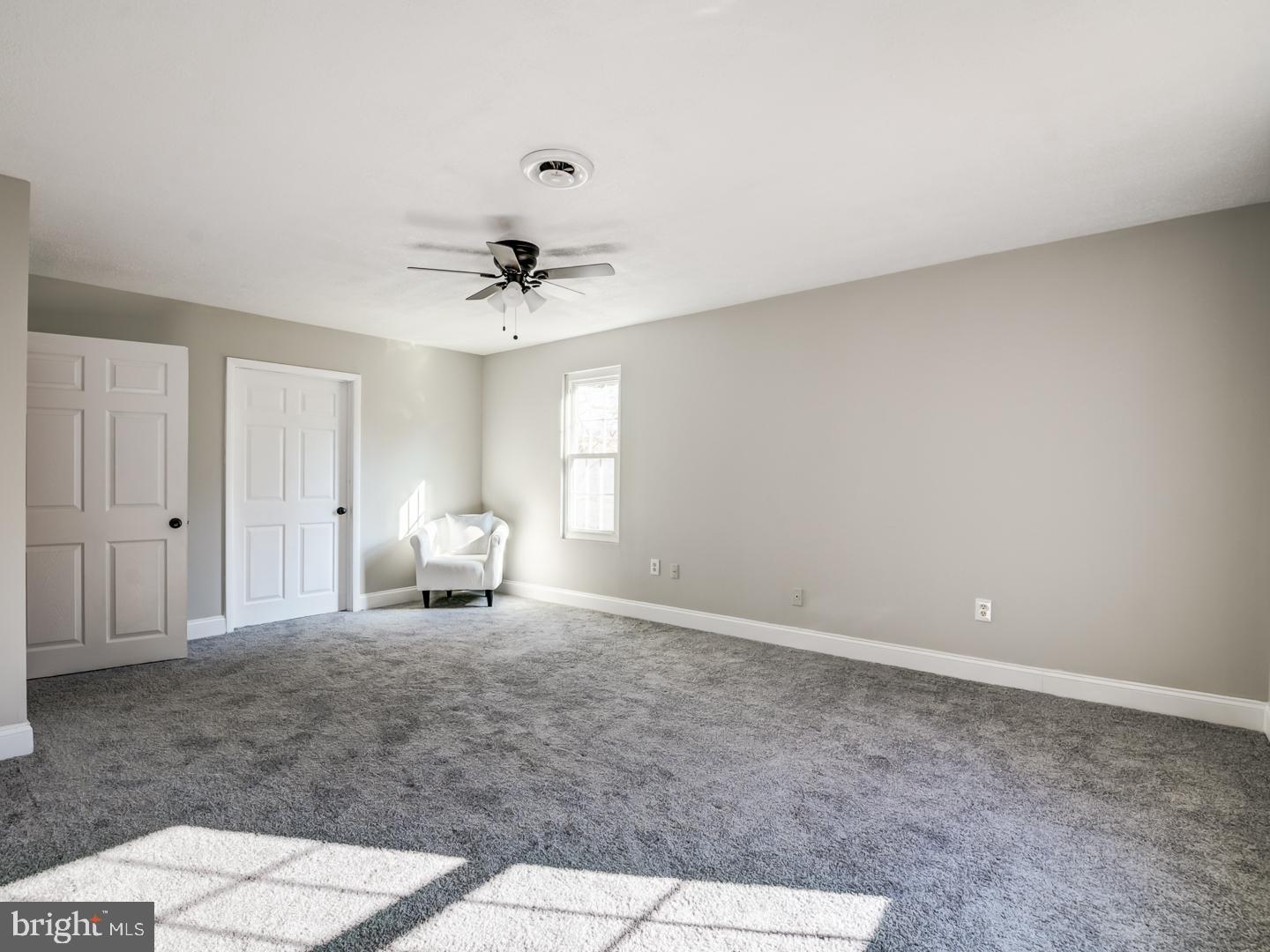 Mostly empty bedroom with gray carpet, ceiling fan, and a white armchair beside the window.