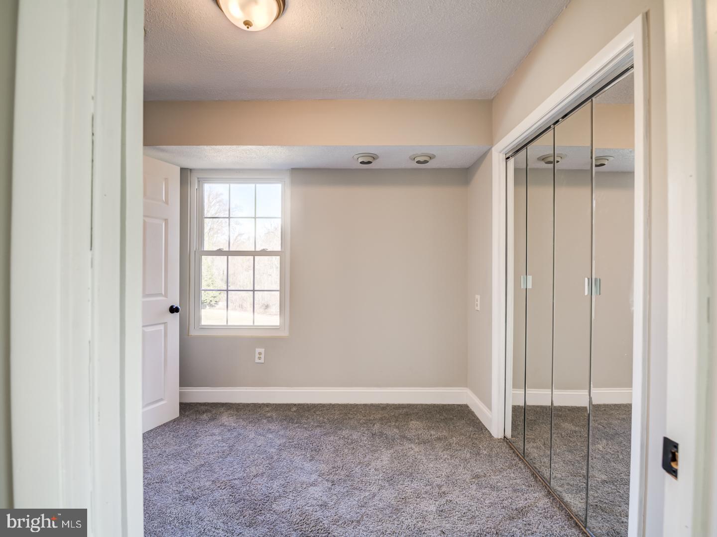 Empty bedroom with gray carpet, a window, and mirrored sliding closet doors.