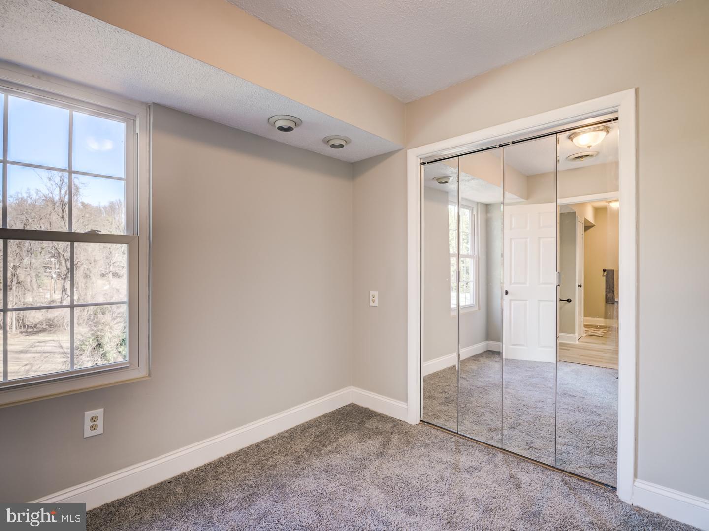 Empty carpeted bedroom with a large window and mirrored sliding closet doors.