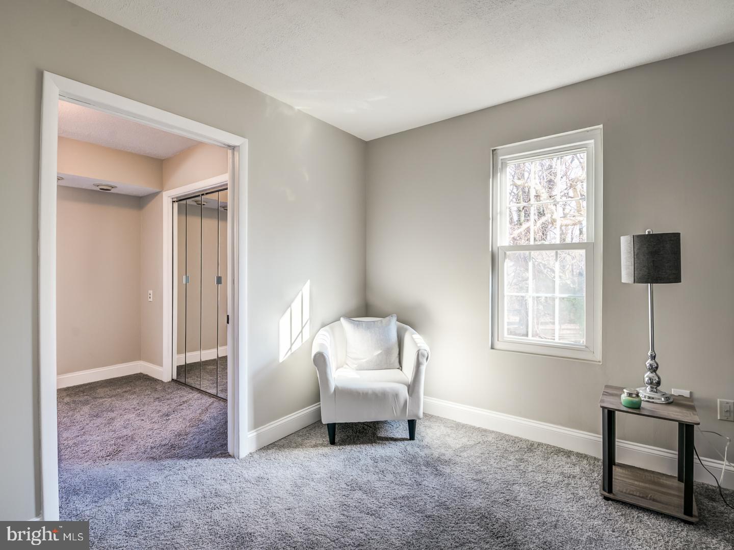 Carpeted bedroom with mirrored closet doors, a white armchair, and a sunlit window.