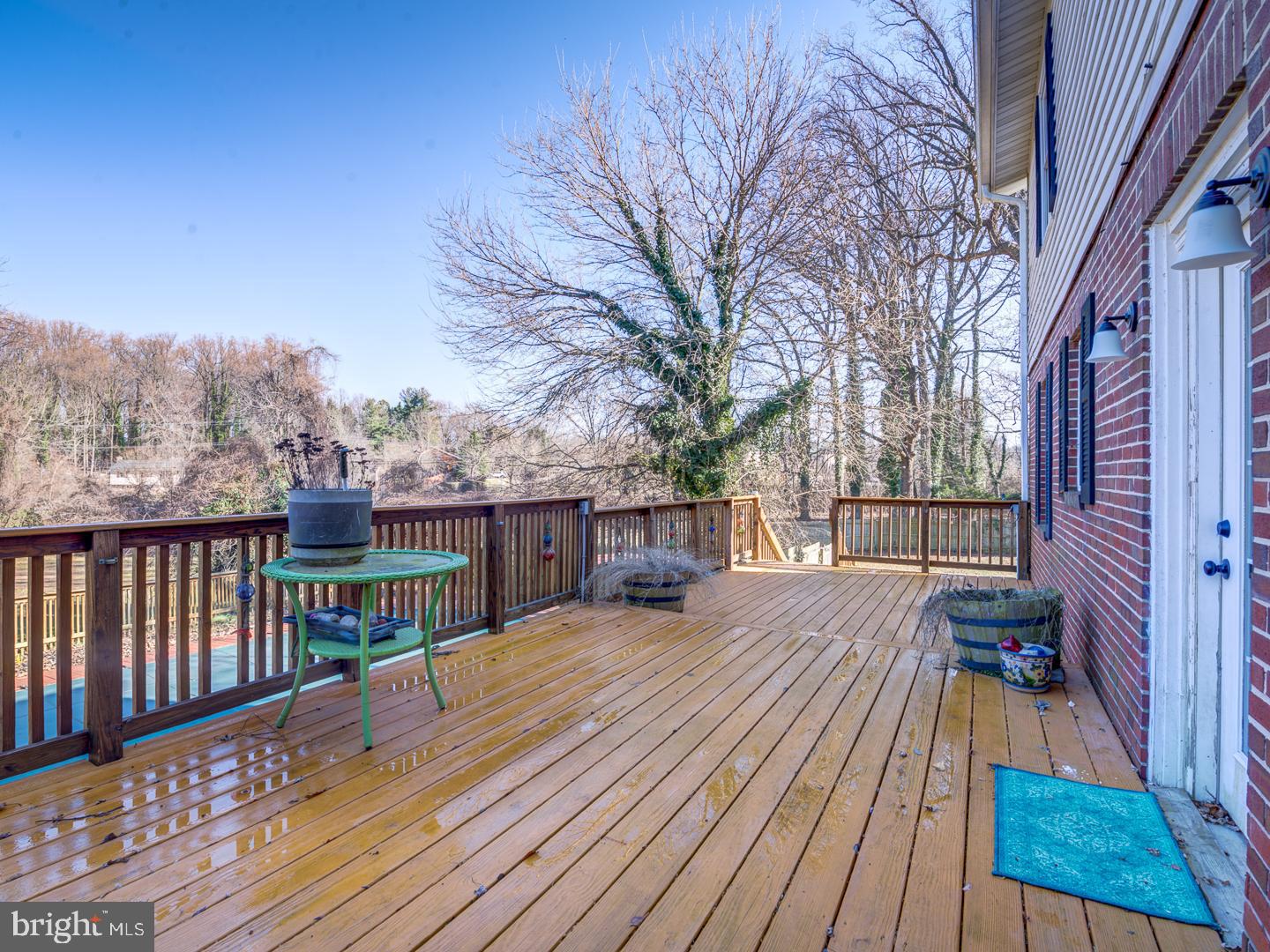 Wooden deck beside a brick house, with a green table and leafless trees beyond the railing.