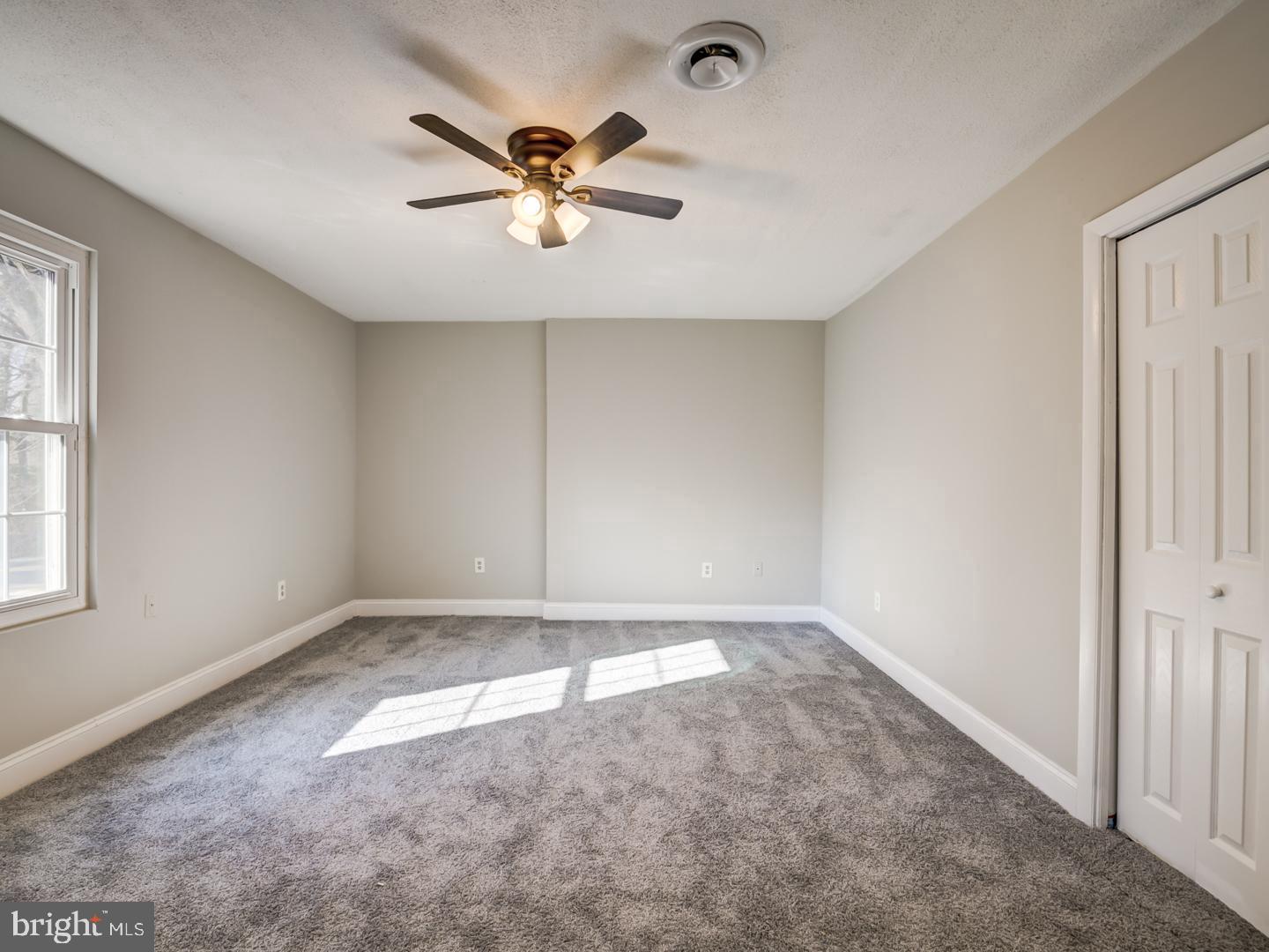Empty bedroom with gray carpet, a ceiling fan, and sunlight streaming through the window.