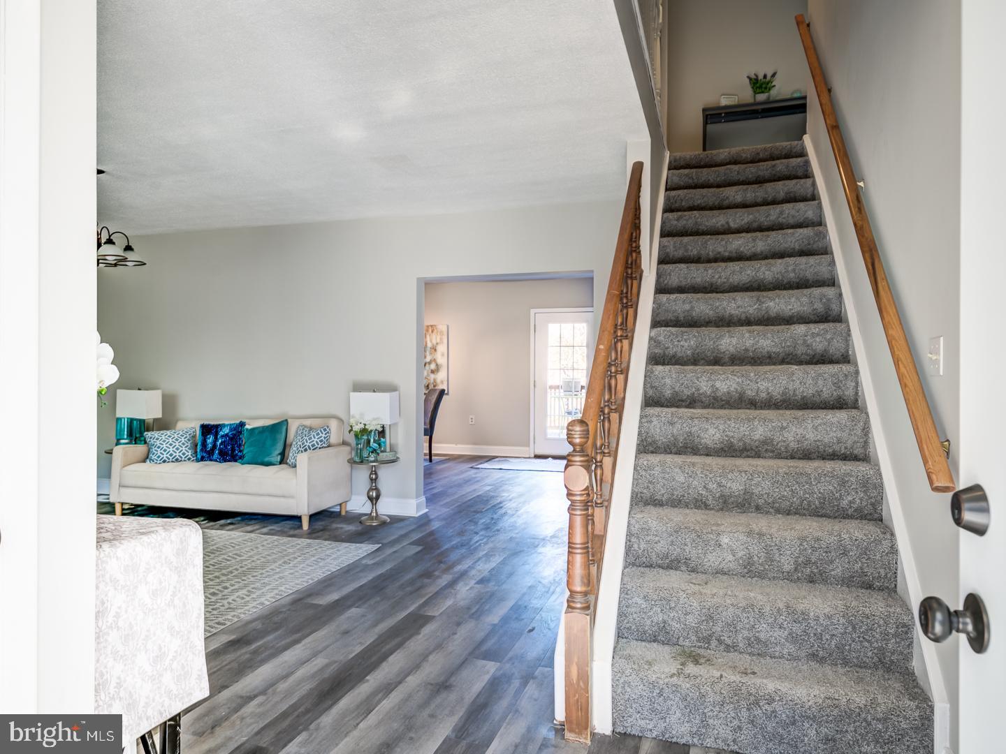 Living room and entryway with a carpeted staircase, wood handrails, and a cream sofa with blue pillows.