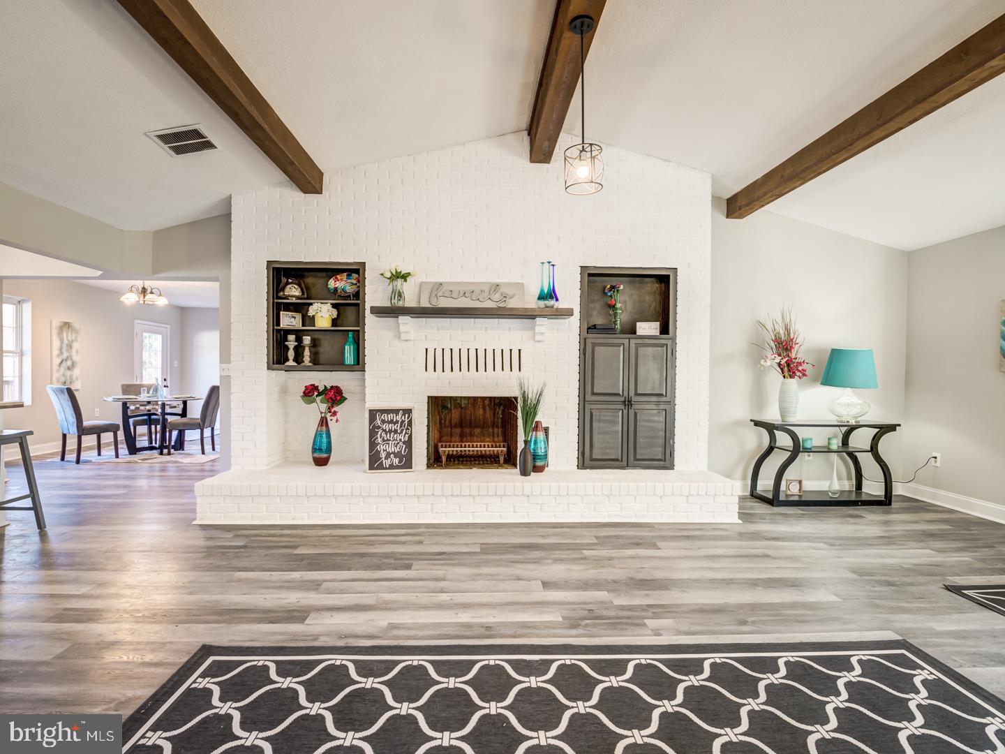 Living room with a white brick fireplace, dark built-ins, exposed ceiling beams, and gray wood-look floors.