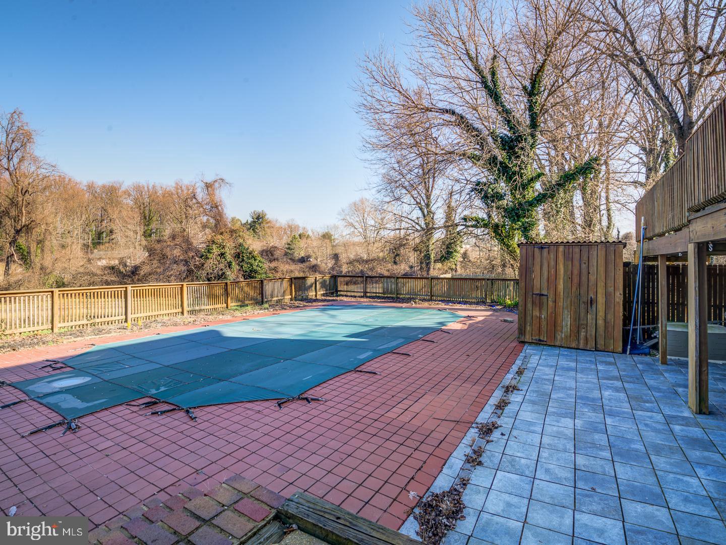 Covered backyard pool surrounded by brick pavers and a tiled patio, with a wooden fence and leafless trees.