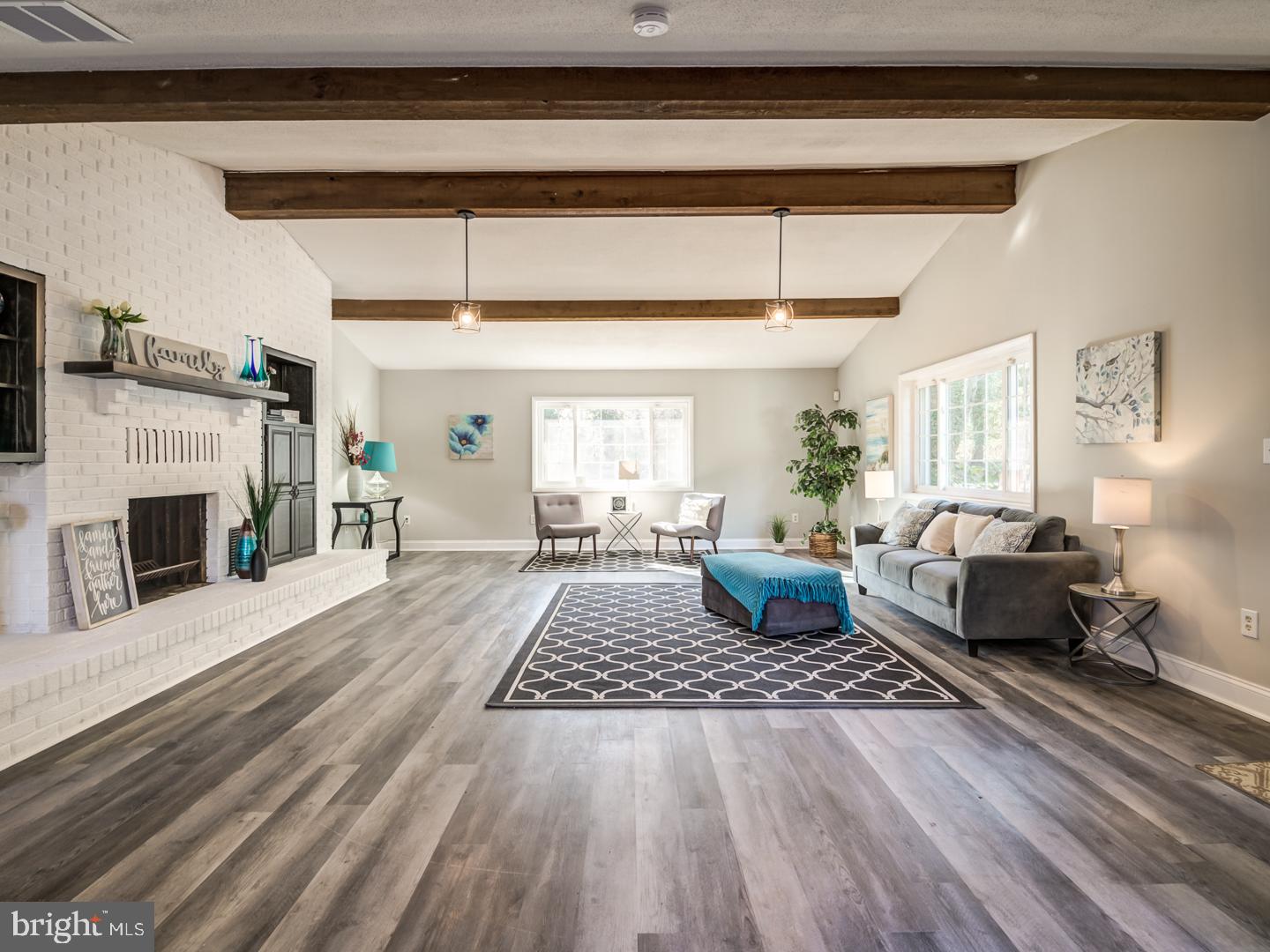 Living room with a white brick fireplace, exposed wood beams, and a gray sofa beside large windows.
