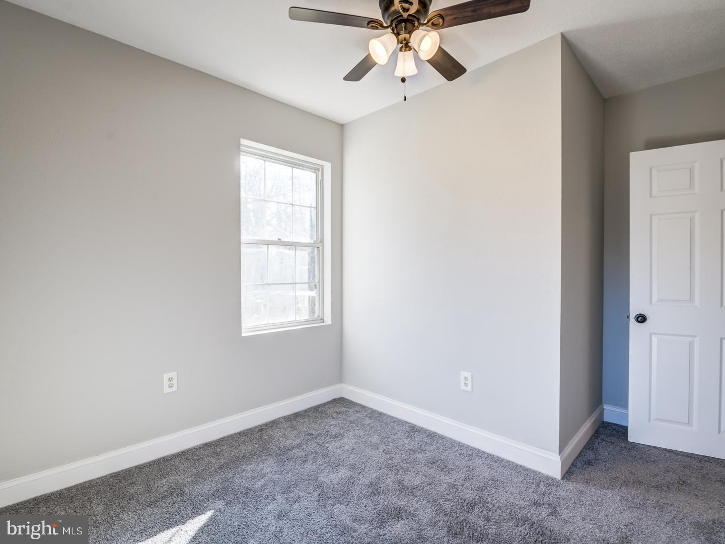 Empty bedroom with gray carpet, a bright window, and a ceiling fan with lights.