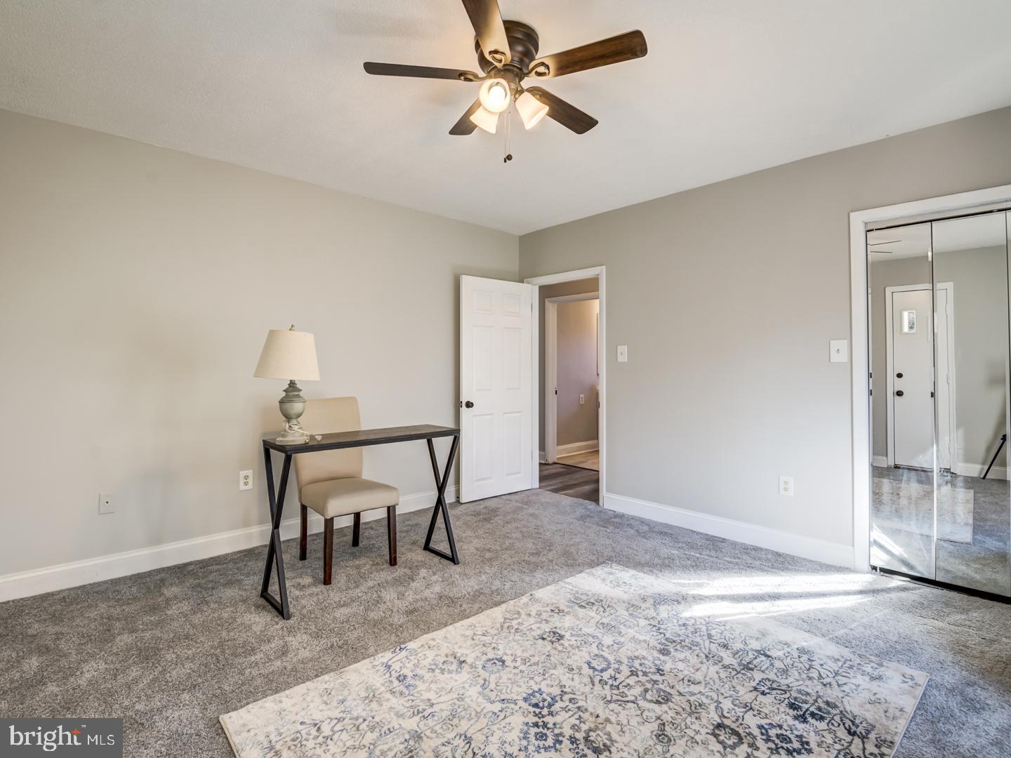 Carpeted bedroom with mirrored closet doors, a ceiling fan, and a small desk and chair.