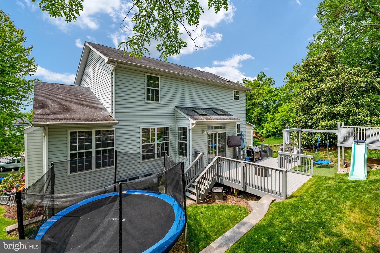 Rear exterior of a two-story home with a raised deck, netted trampoline, and children's playset with a slide.