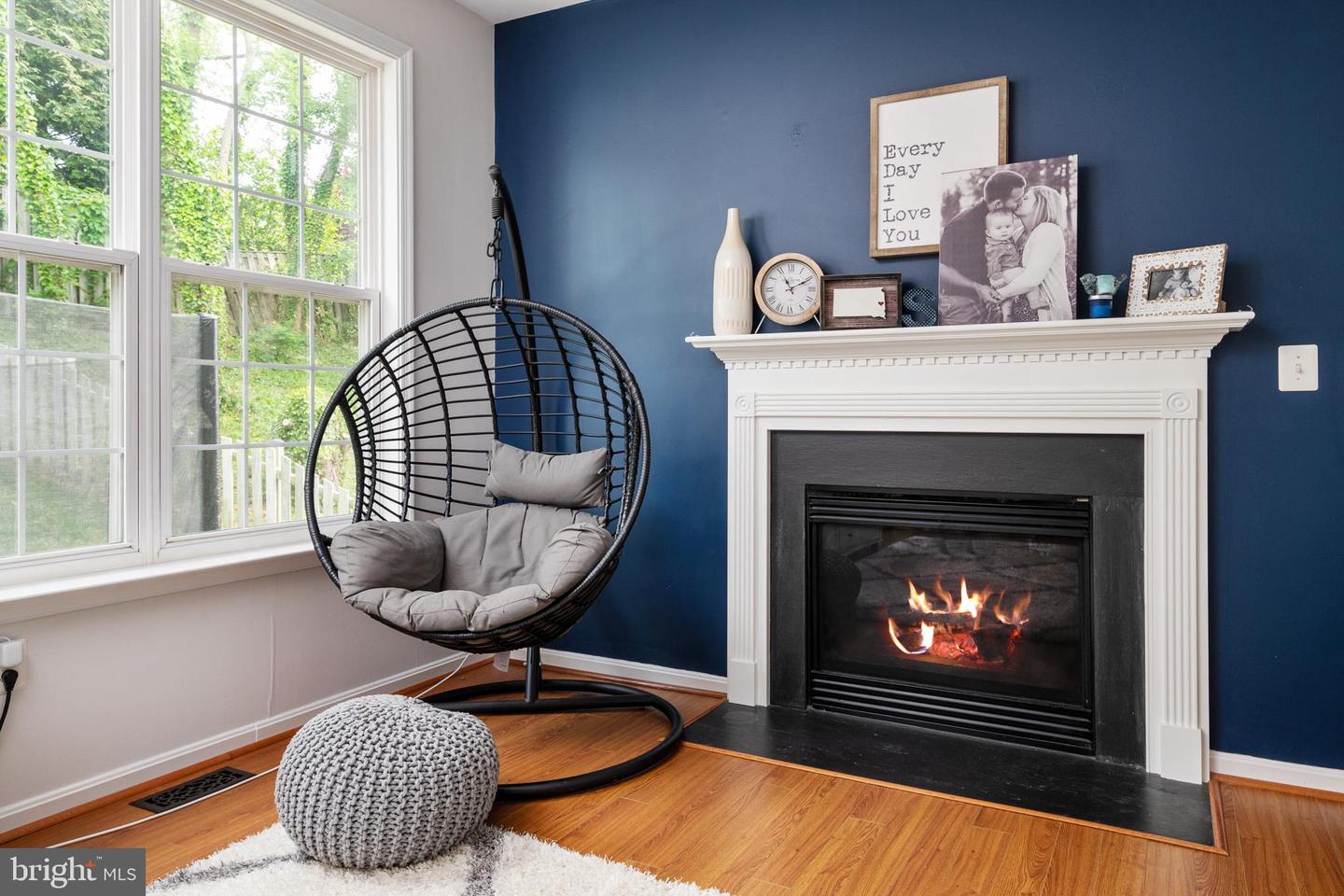 Living room with a lit fireplace, hanging chair, and large windows overlooking greenery.