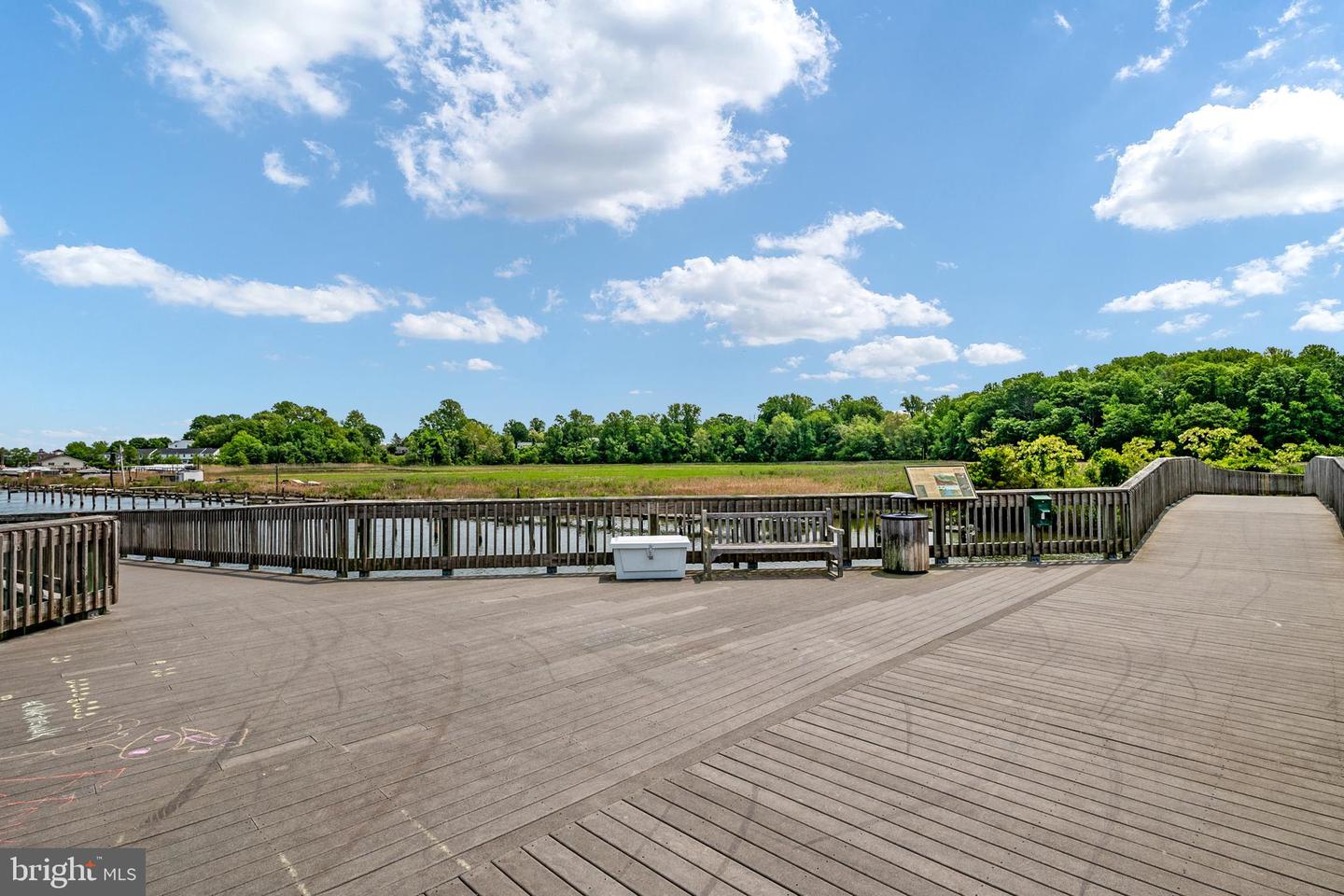 Waterfront boardwalk and deck beside marshland, with a wooded shoreline in the distance.