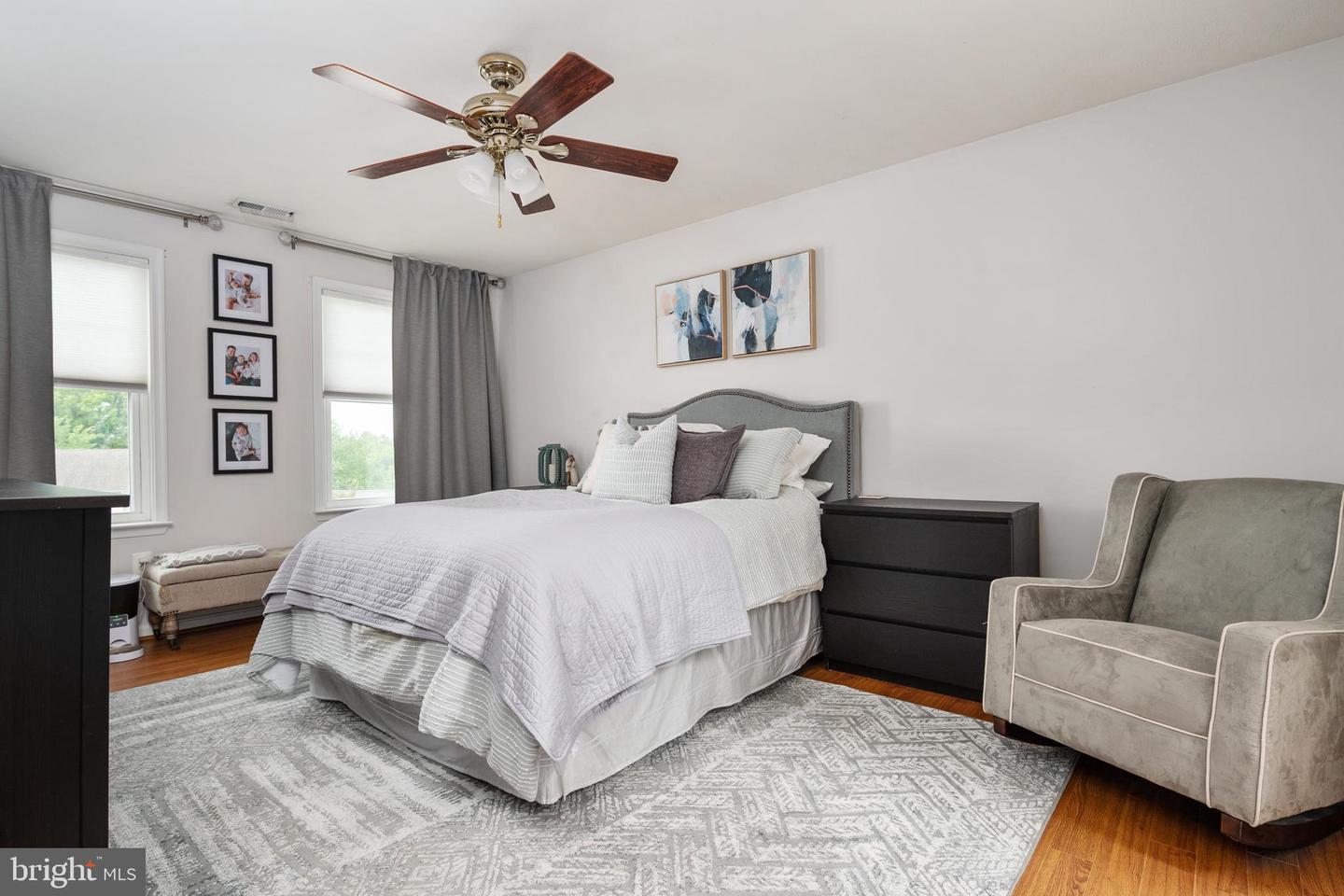 Furnished bedroom with a white bed, patterned rug, ceiling fan, and gray armchair.
