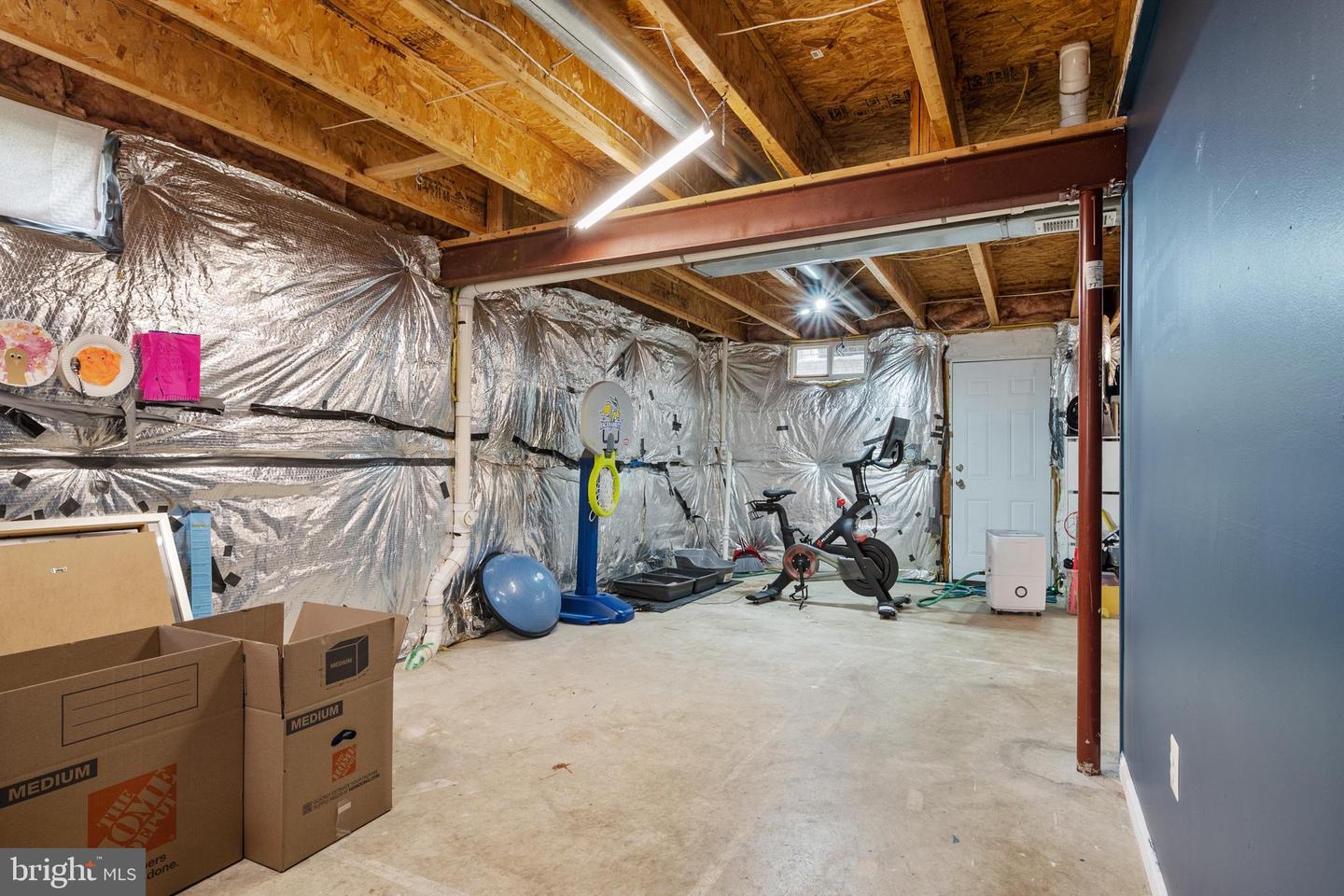 Unfinished basement with exposed ceiling framing, foil-covered walls, exercise equipment, and moving boxes.
