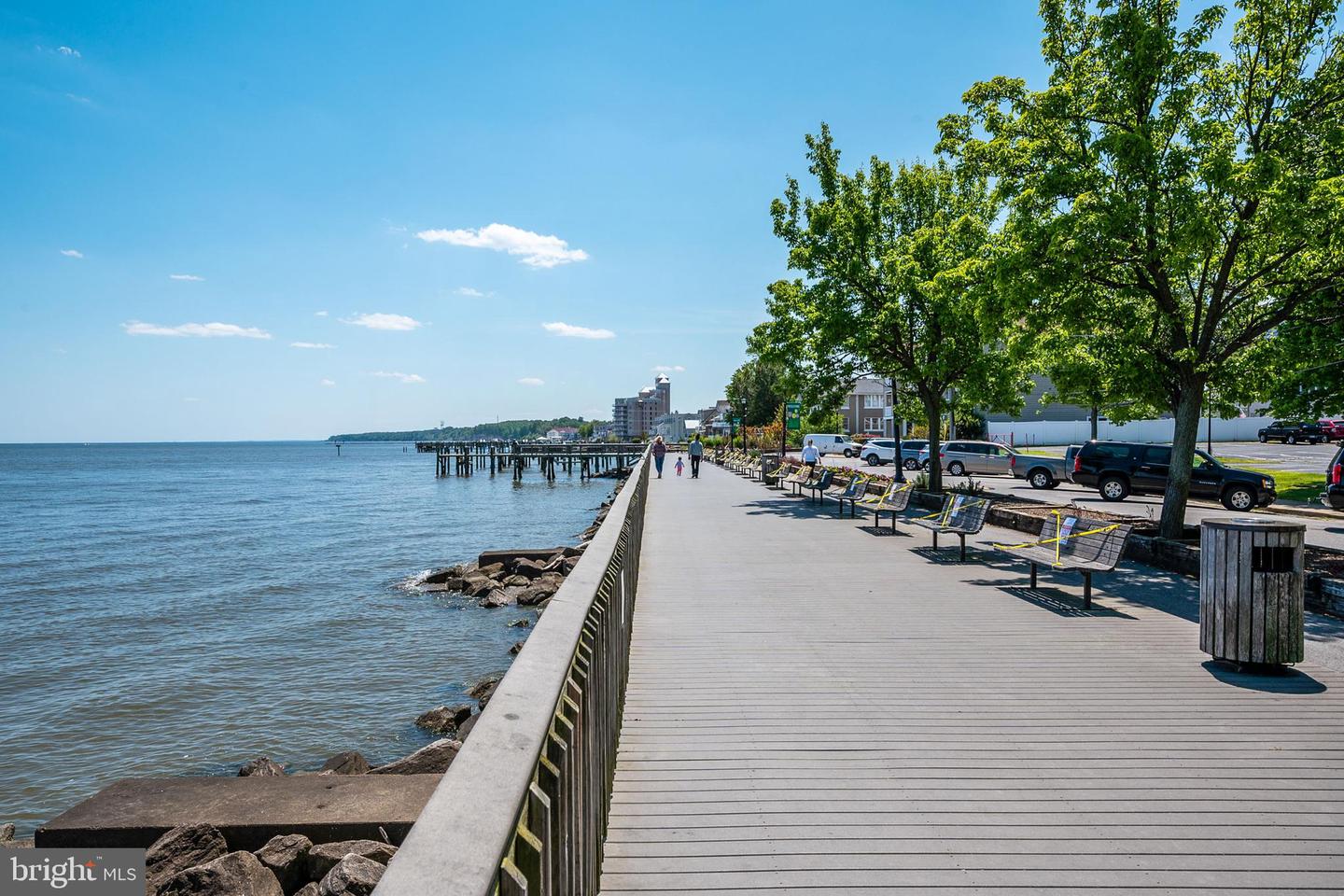Waterfront boardwalk with shaded benches, calm water, and a pier in the distance.