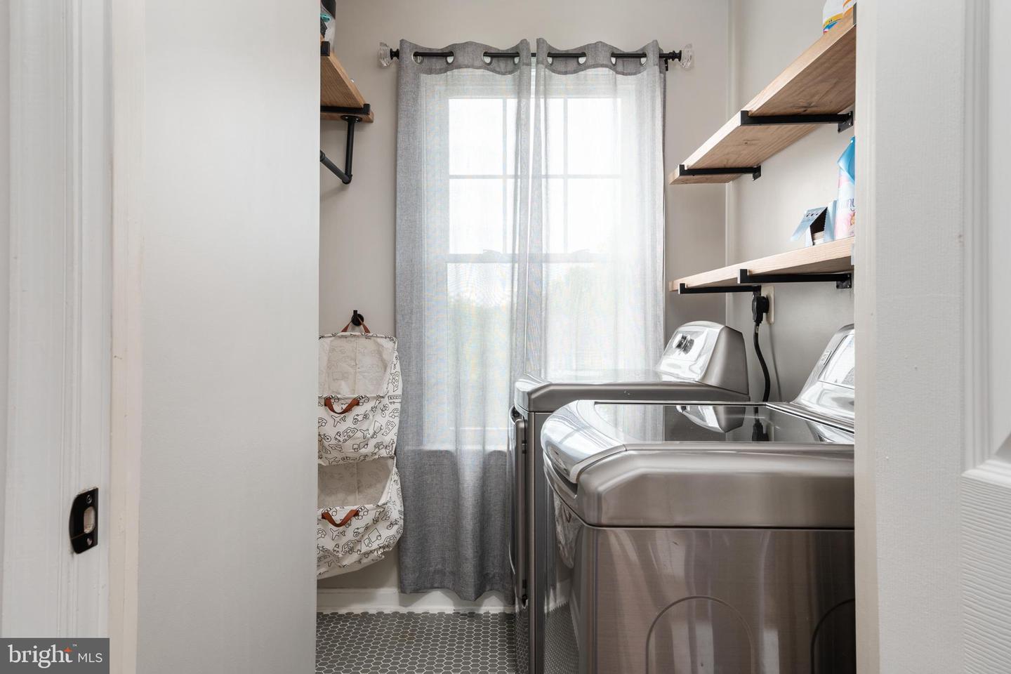 Laundry room with stainless steel washer and dryer, open wood shelves, and a curtained window.