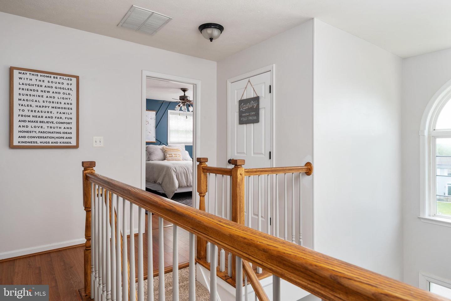 Upstairs landing with a wooden railing, arched window, and view into a blue bedroom.