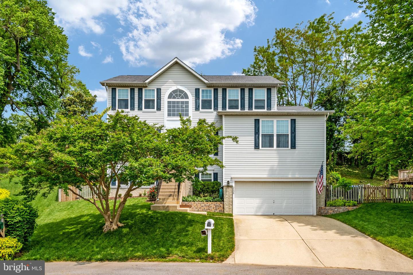 Two-story gray house with dark shutters, a white garage, and a tree-shaded front lawn.