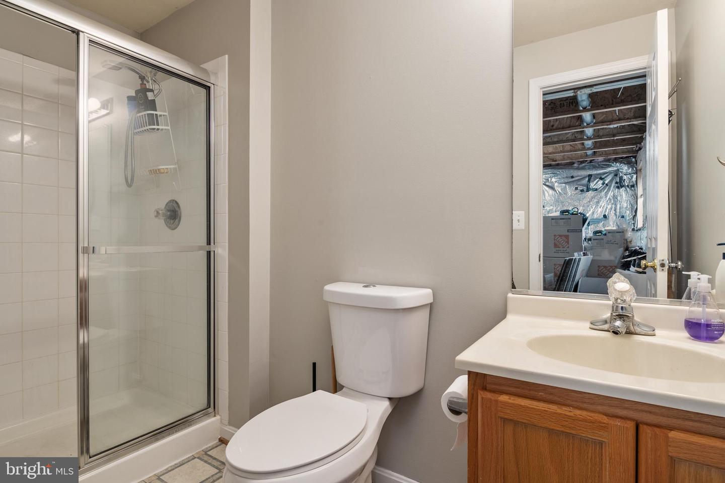 Bathroom with glass shower, toilet, and wood vanity; open doorway reveals an unfinished adjacent room.