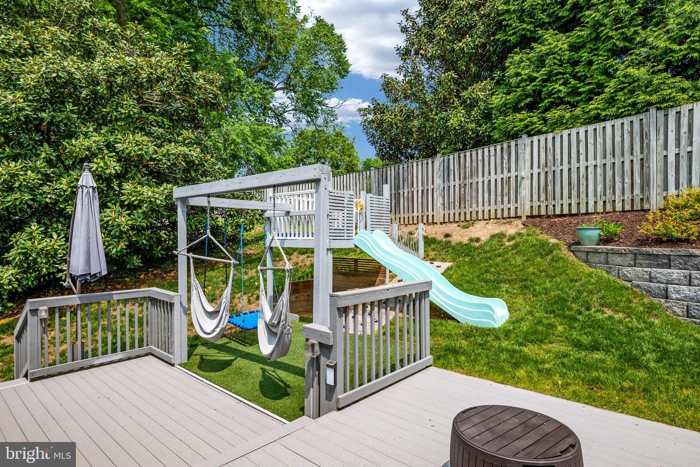 Backyard play area with a wooden swing set, aqua slide, and fenced deck.