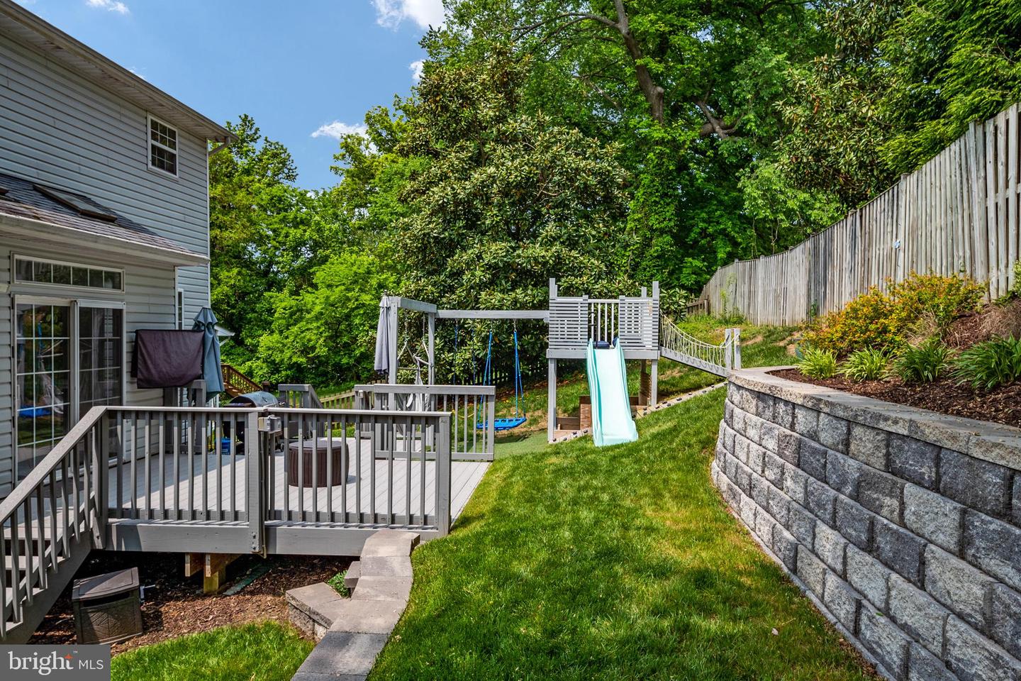 Backyard with a raised deck, children's playset, stone retaining wall, and wooden fence.