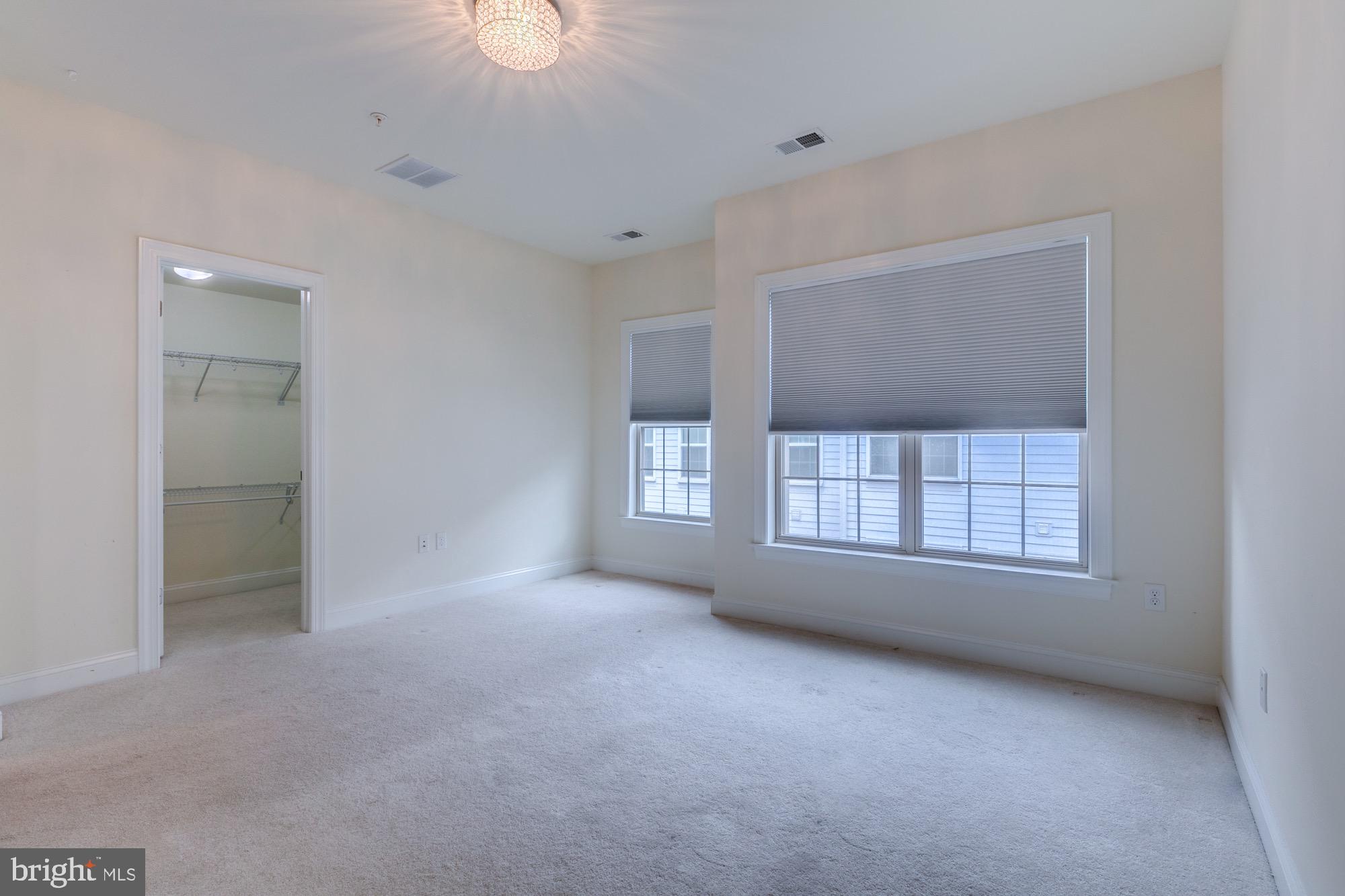 Empty carpeted bedroom with a walk-in closet, two windows with blinds, and a decorative ceiling light.
