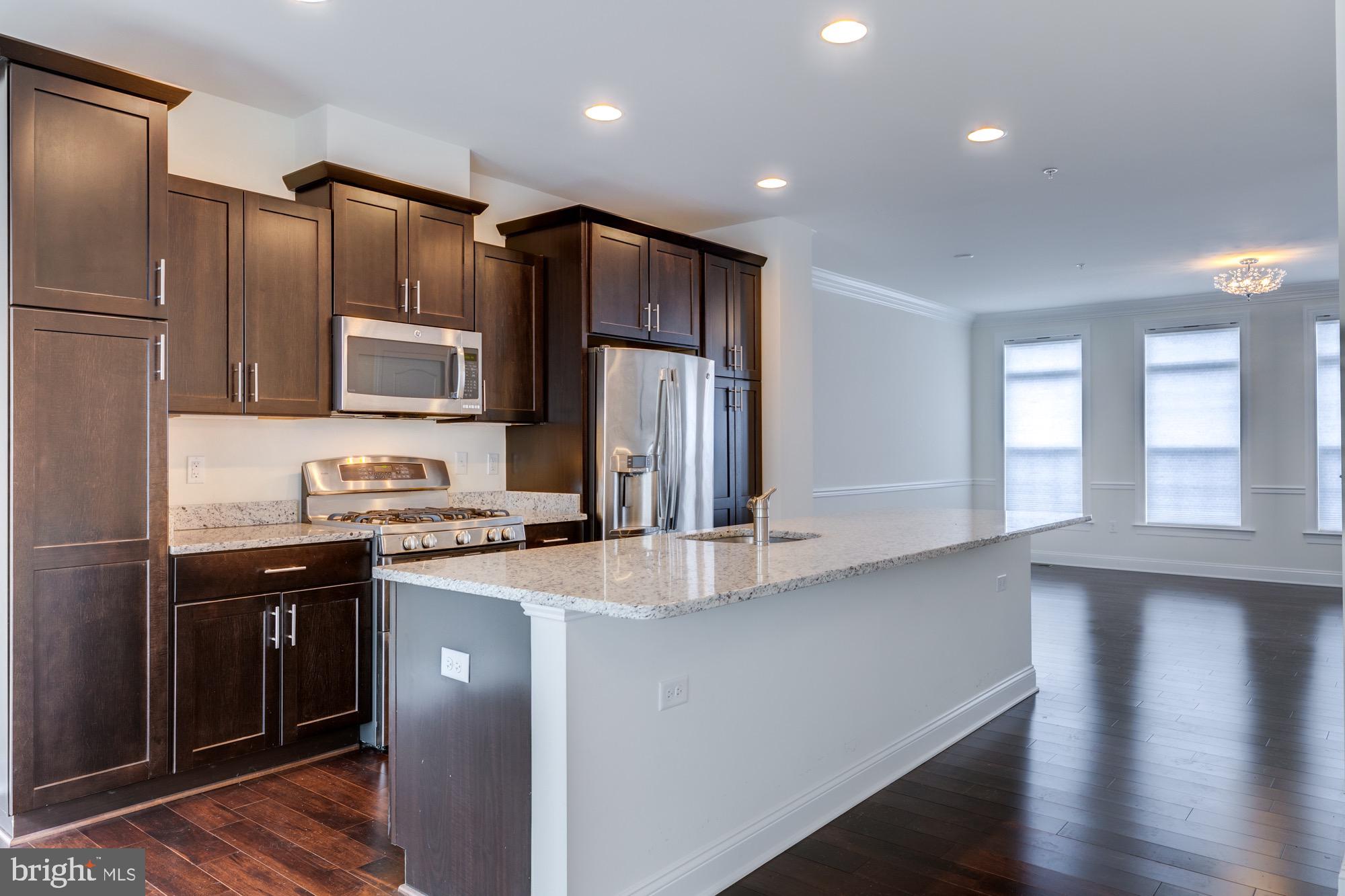 Kitchen with dark wood cabinets, stainless steel appliances, and a large granite island opening onto a bright living area.