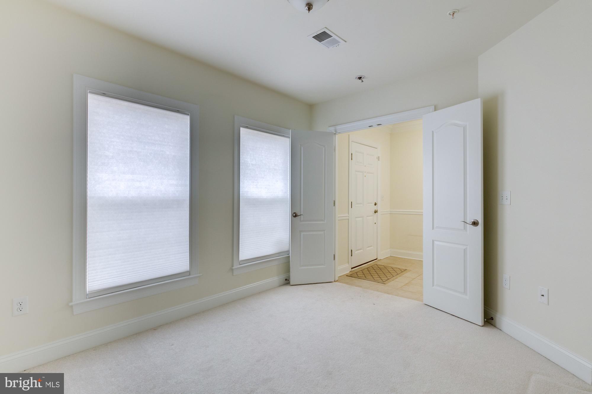 Empty carpeted room with two shaded windows and open doors to a tiled entry hall.