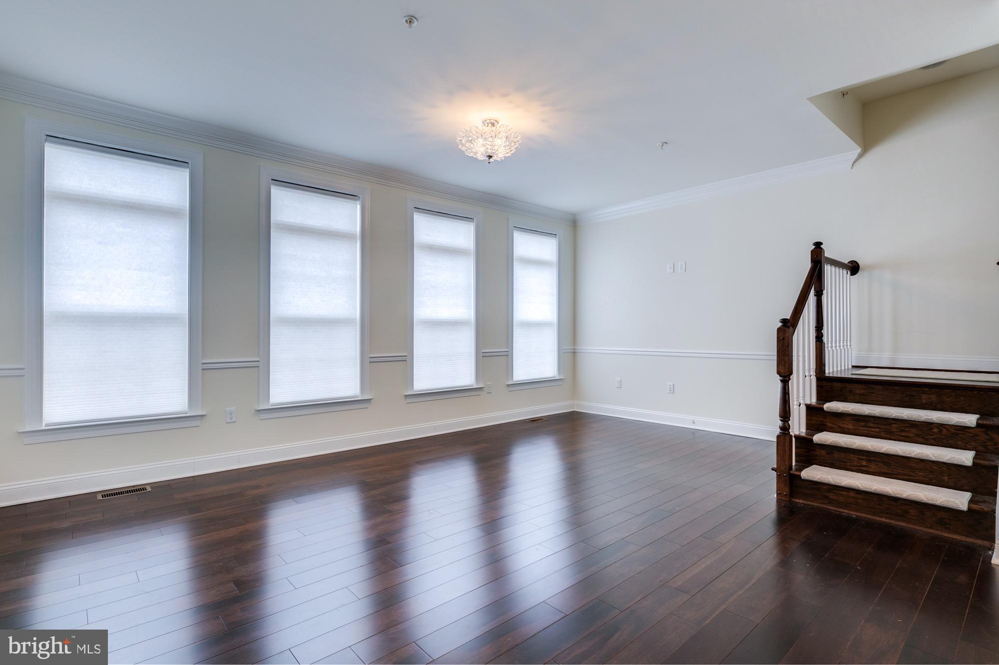 Empty room with dark hardwood floors, four tall shaded windows, a chandelier, and a dark wood staircase.