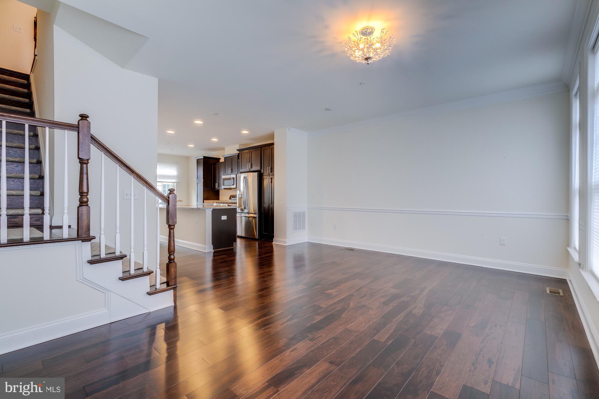 Open living area with dark hardwood floors, staircase, and adjoining kitchen with stainless steel appliances.