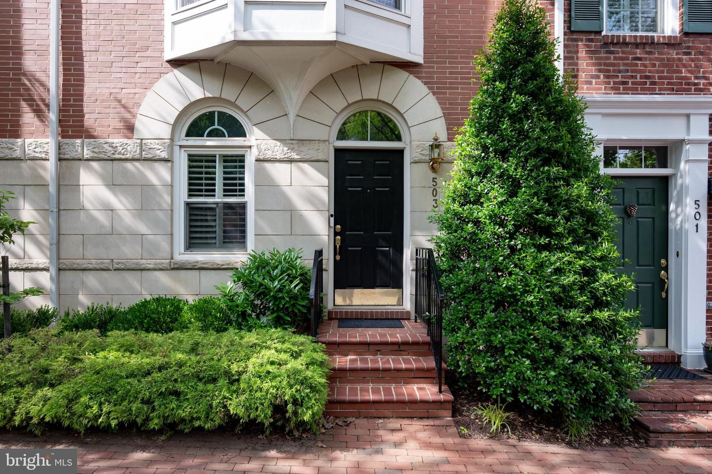 Brick home entrance with arched stone surround, black door, brick steps, and mature landscaping.