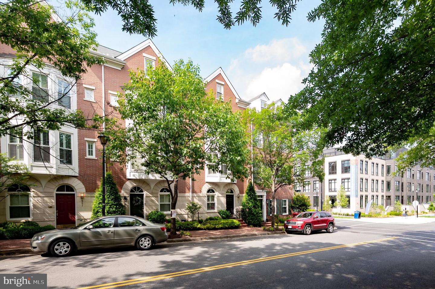Red-brick townhouses with arched entrances beneath leafy trees along a city street.
