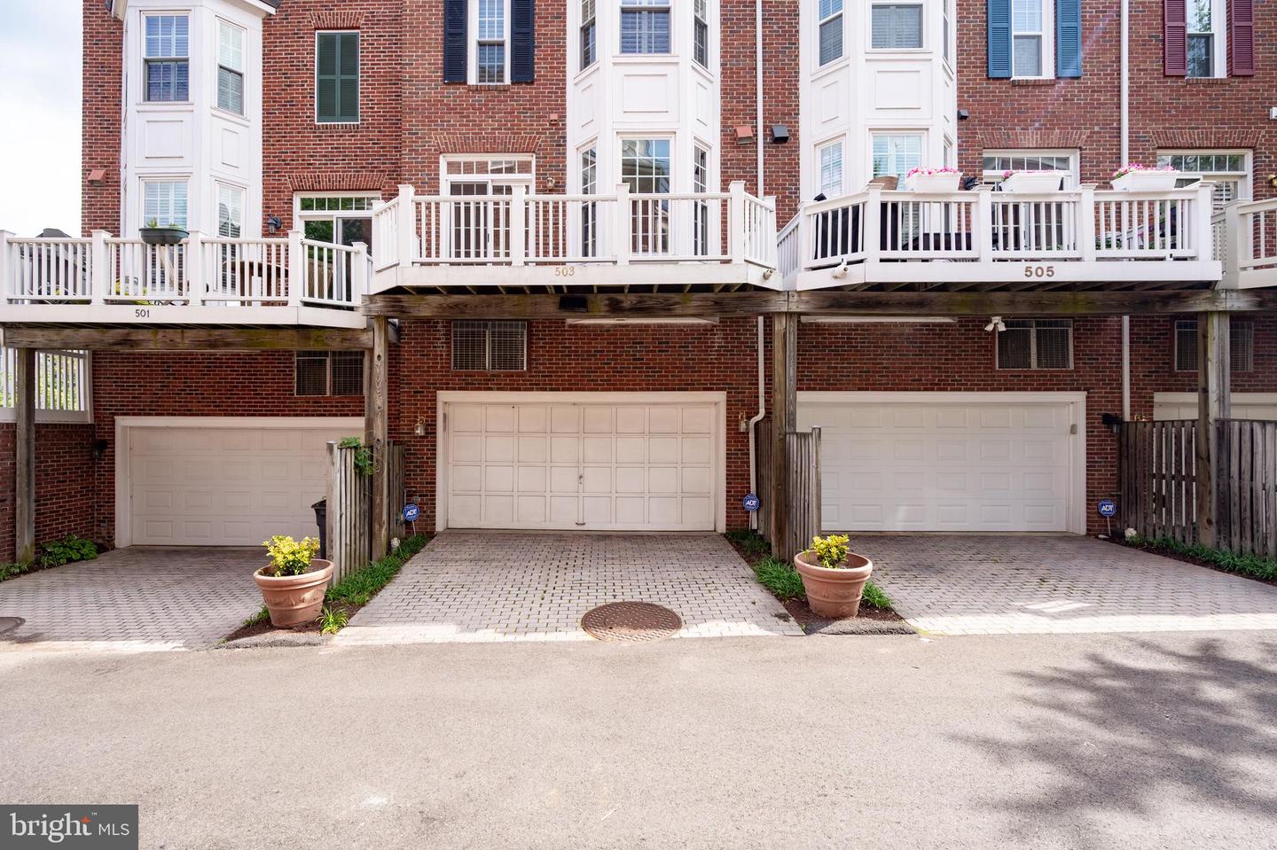 Rear exterior of brick townhouses with elevated white decks, garage doors, and paved driveways.