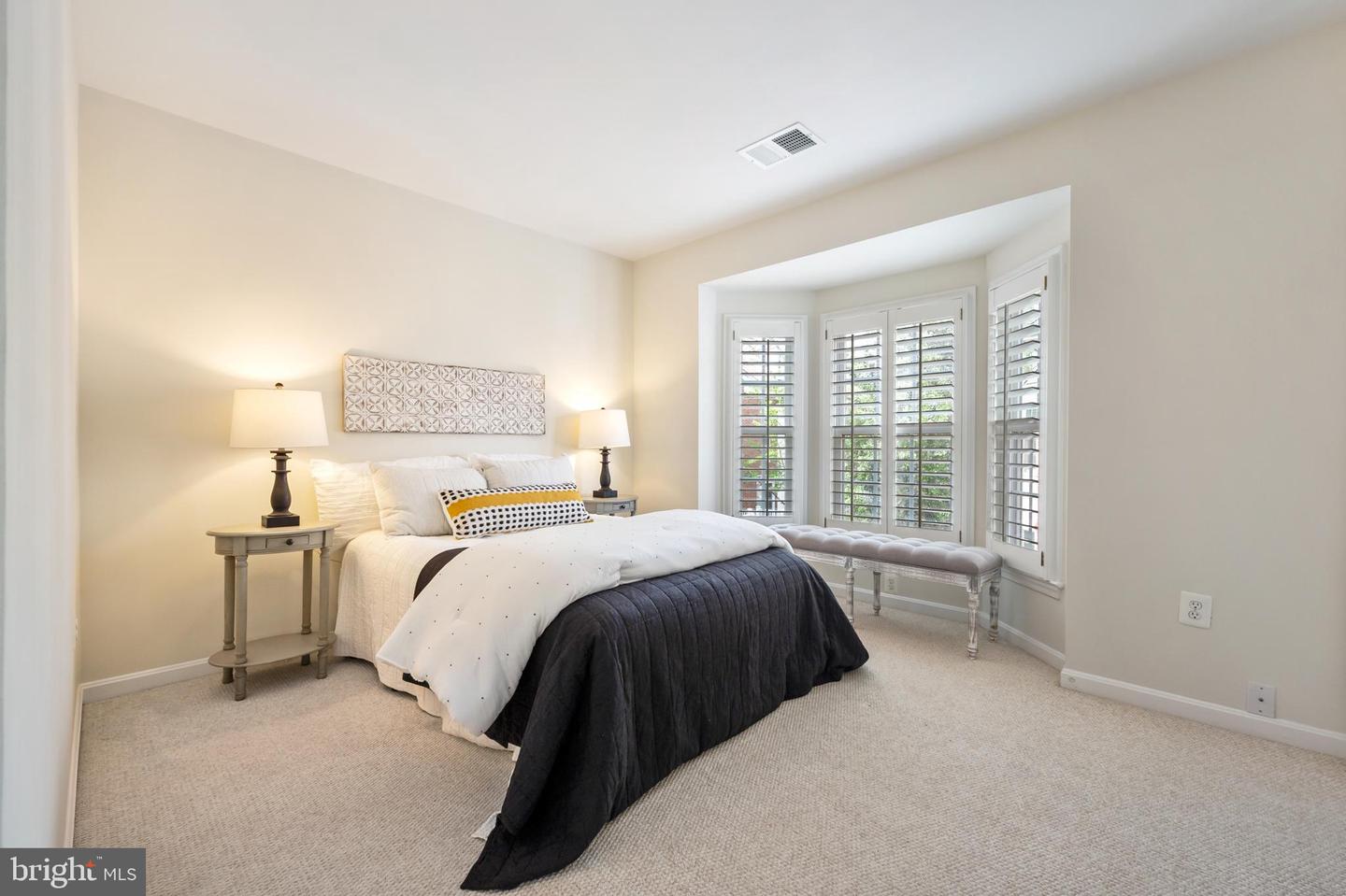 Staged bedroom with a large bed, matching bedside lamps, and plantation shutters on a bay window.